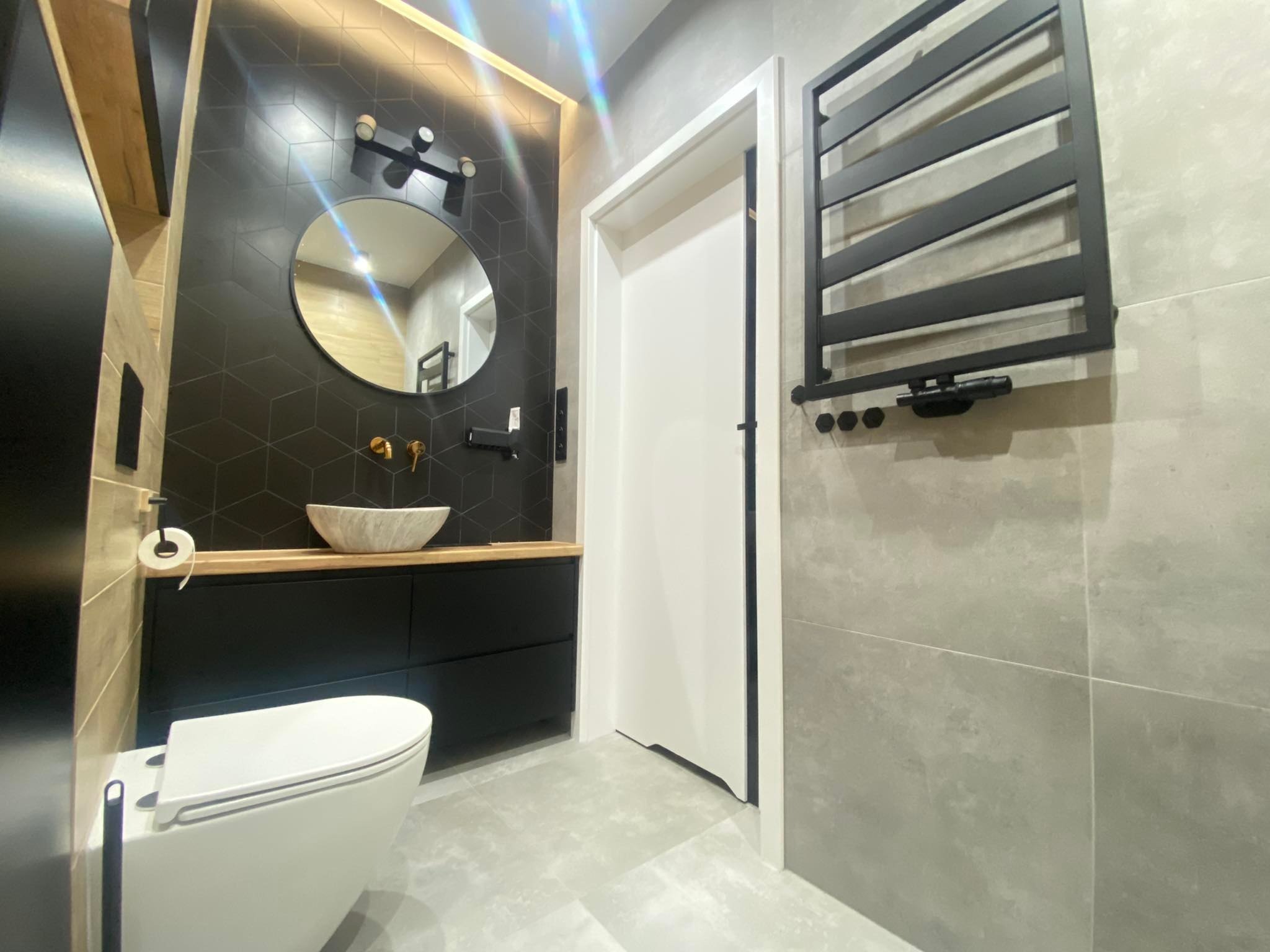 Nowoczesna łazienka z czarnymi płytkami heksagonalnymi, okrągłym lustrem, umywalką nablatową w kształcie muszli, czarną szafką pod umywalką z drewnianym blatem, wiszącą toaletą i grzejnikiem...
