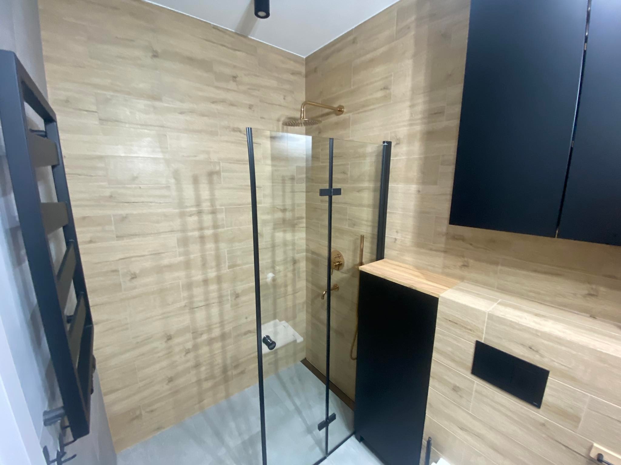 Nowoczesna łazienka z kabiną prysznicową typu walk-in, złotą armaturą i płytkami imitującymi drewno na ścianach, uzupełniona czarnymi elementami wyposażenia.