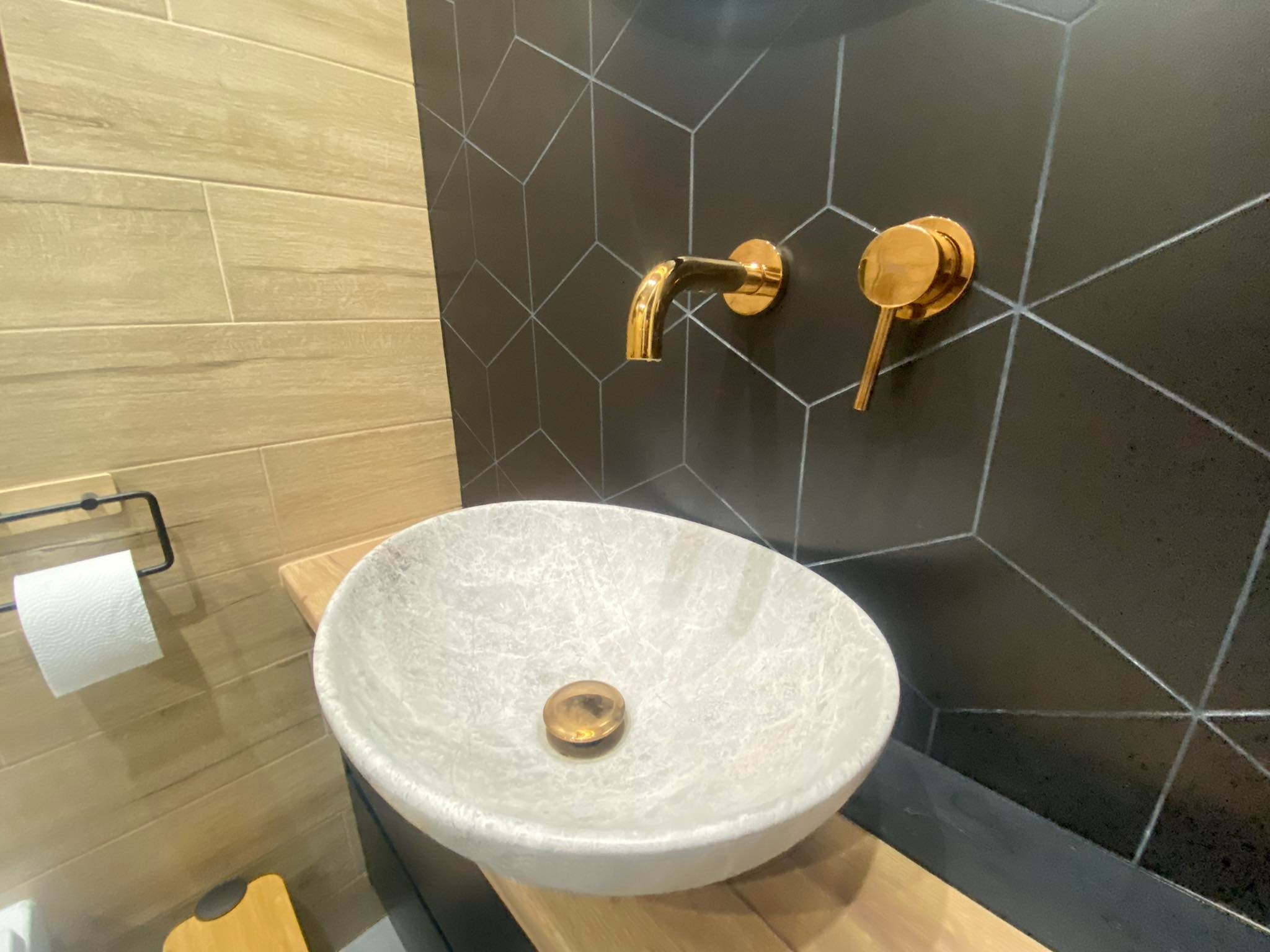 Nowoczesna łazienka z umywalką nablatową w kolorze jasnego marmuru, złotą armaturą ścienną i czarną ścianą z geometrycznym wzorem, drewniany blat i akcesoria.