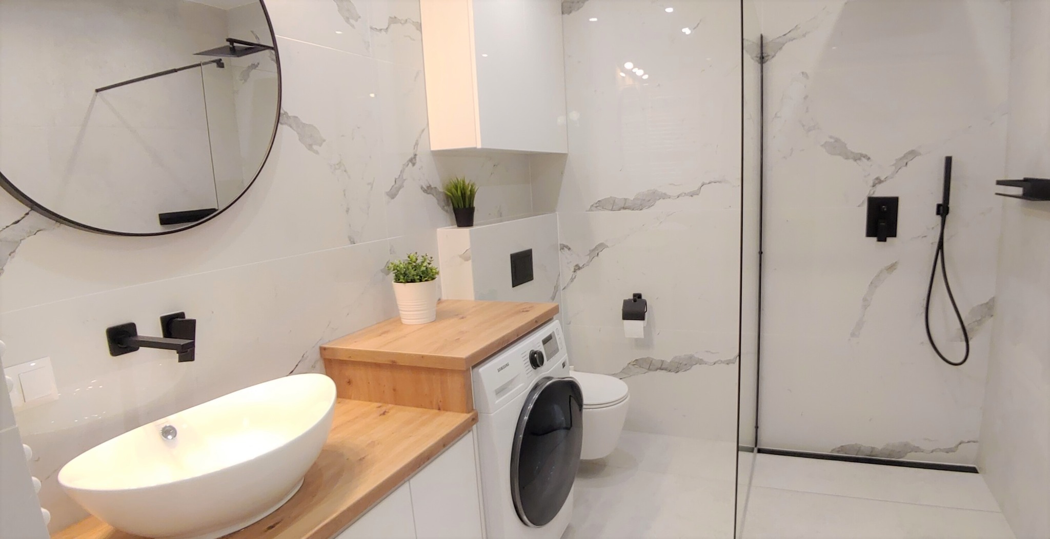 Nowoczesna łazienka w bieli z marmurowym wzorem na ścianach, wyposażona w okrągłe lustro z czarną ramą, owalną umywalkę nablatową, drewniany blat, pralkę, toaletę i kabinę prysznicową z czarnymi...