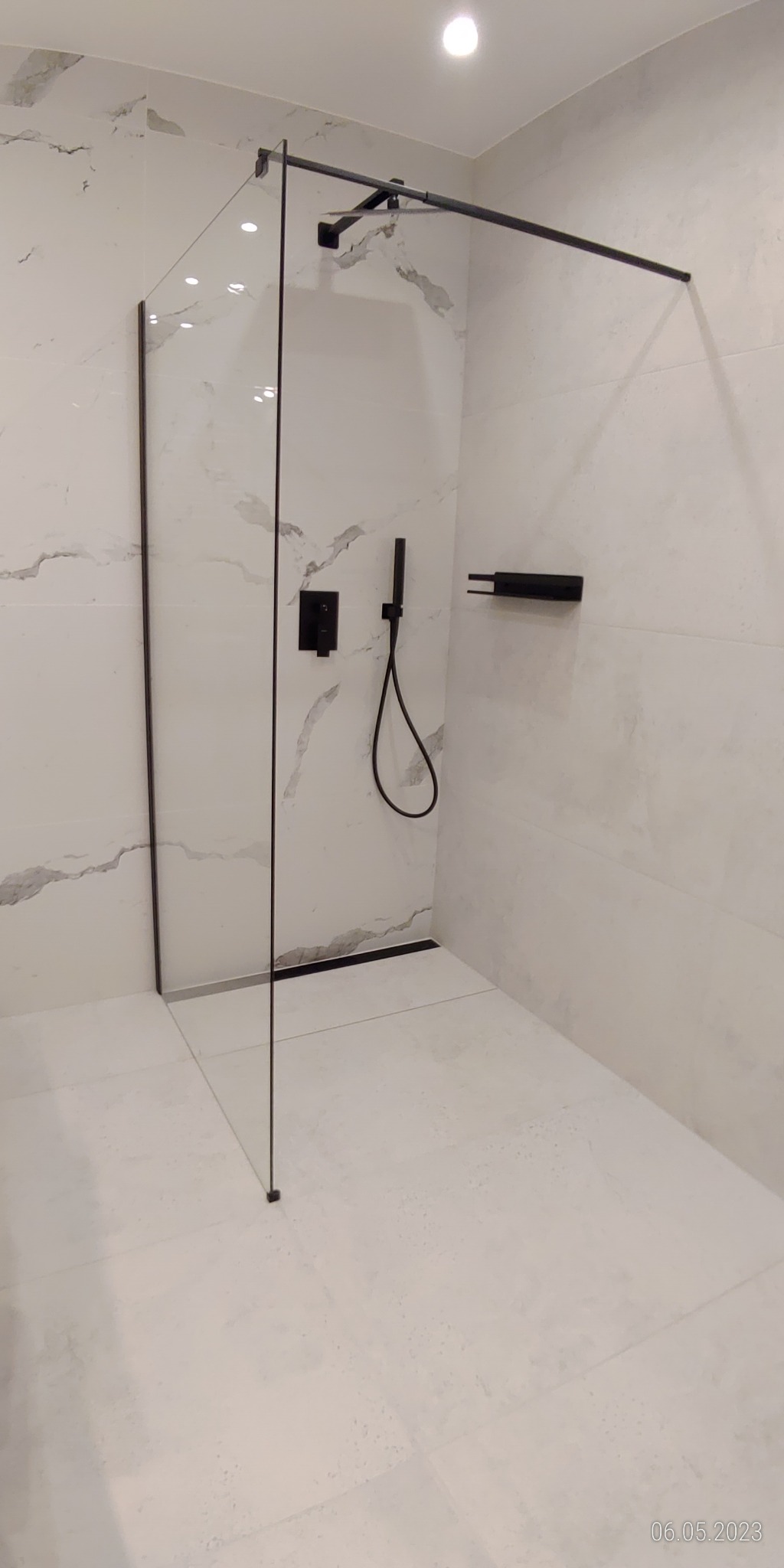 Nowoczesna kabina prysznicowa typu walk-in z czarnymi elementami armatury i odpływem liniowym, wykończona białymi płytkami imitującymi marmur.