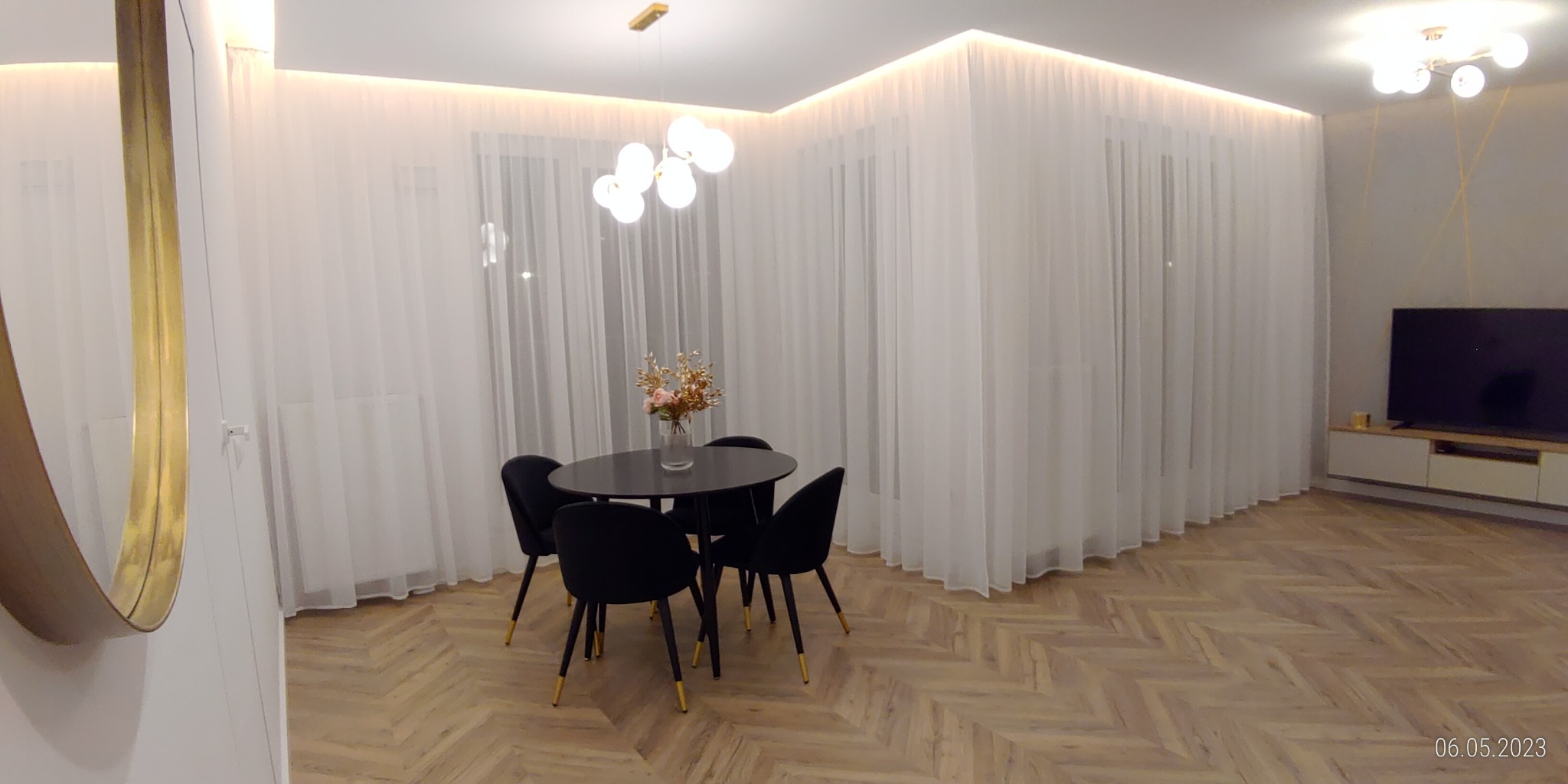 Jasne wnętrze z parkietem w jodełkę, okna z białymi zasłonami, czarny okrągły stół z bukietem kwiatów i cztery krzesła z złotymi nogami, nowoczesna lampa sufitowa, lustro w złotej ramie.