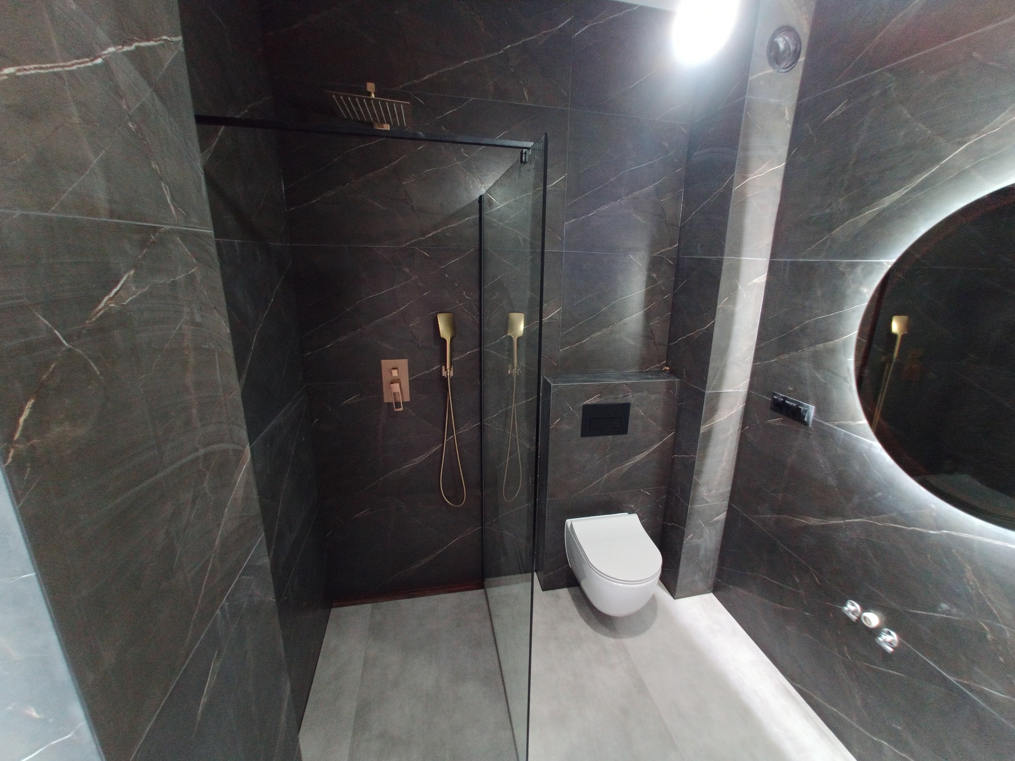 Nowoczesna łazienka z ciemnymi marmurkowymi płytkami, złotą armaturą prysznicową i podświetlanym okrągłym lustrem.
