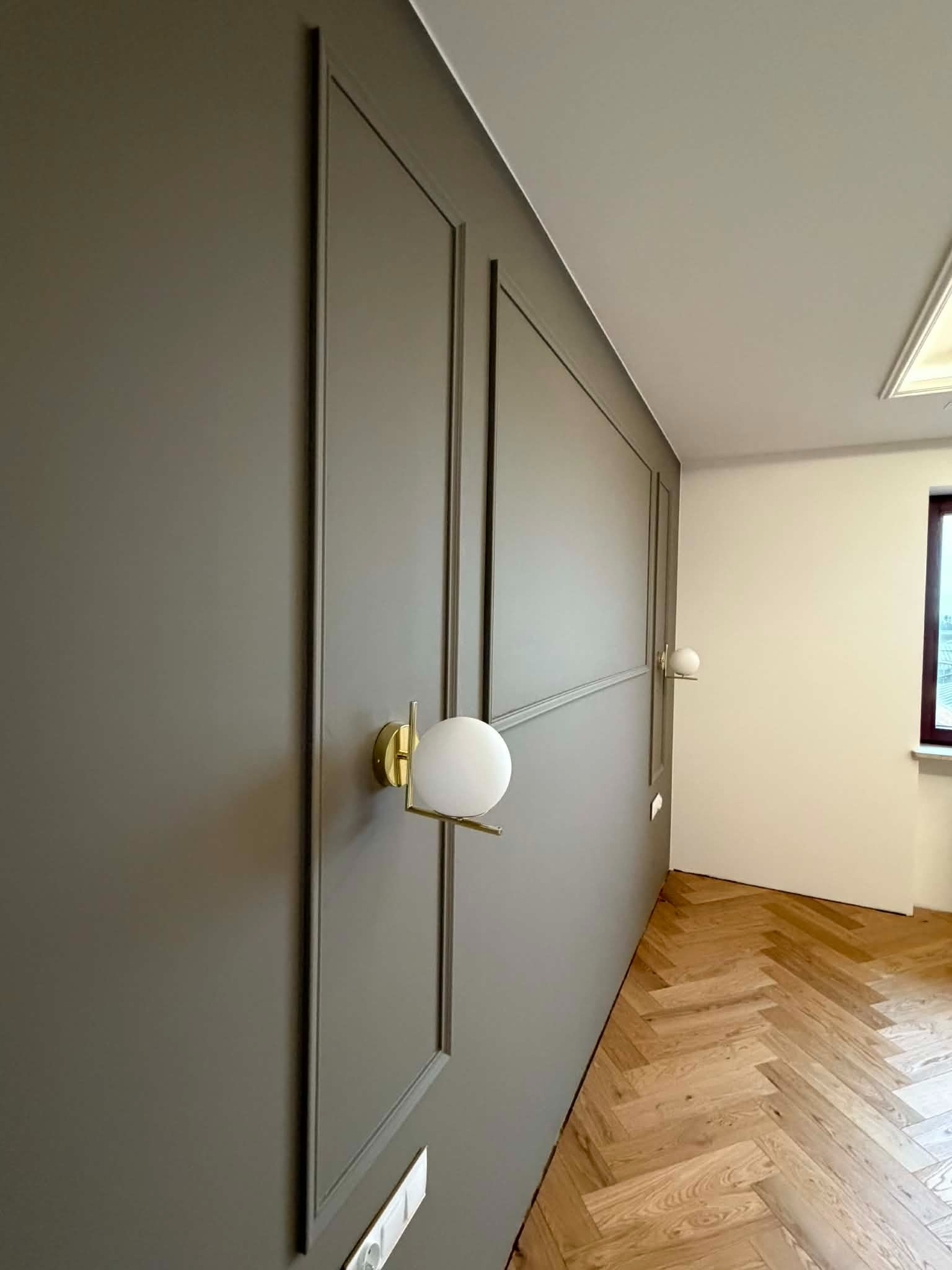 Panelowana ściana w odcieniach szarości z oświetleniem punktowym i parkietem ułożonym w jodełkę. Nowoczesny design wnętrza z dbałością o detale.