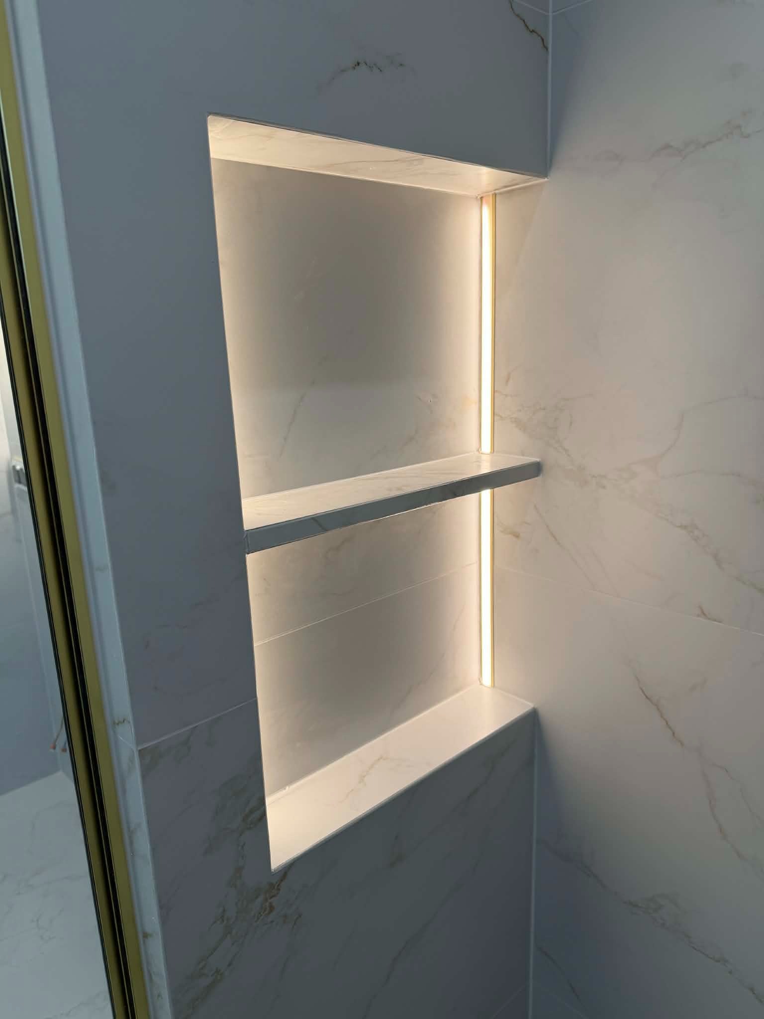 Wnęka z półkami w łazience, wyłożona marmurowymi płytkami, oświetlona ledowym światłem. Minimalistyczny design, nowoczesne wykończenie.