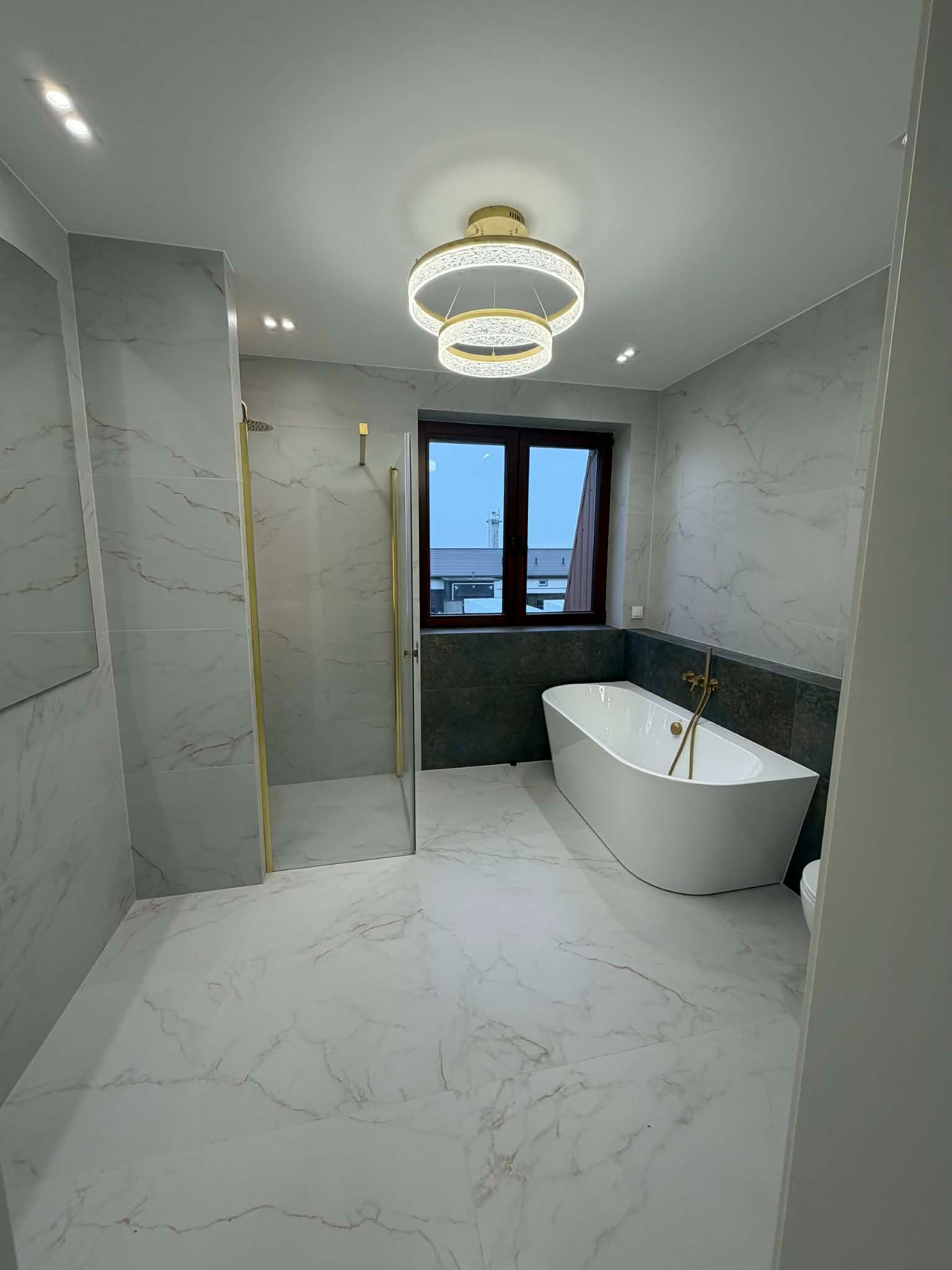 Nowoczesna łazienka z marmurowymi płytkami, kabiną prysznicową ze złotymi detalami i wolnostojącą wanną pod oknem, oświetlona designerską lampą.