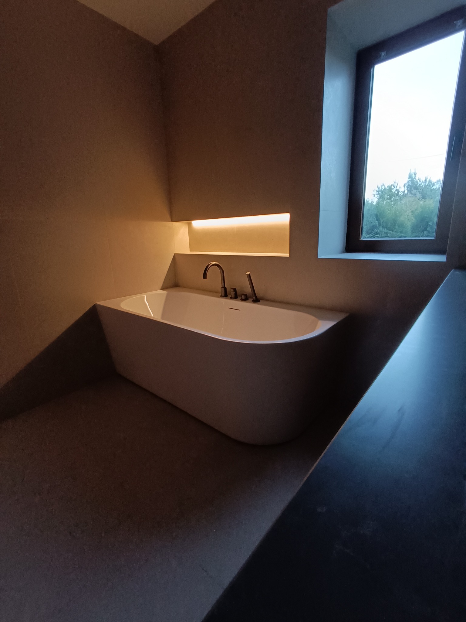 Minimalistyczna łazienka z wolnostojącą wanną podświetloną od góry, okno z widokiem na zieleń. Stonowana kolorystyka, nowoczesny design.