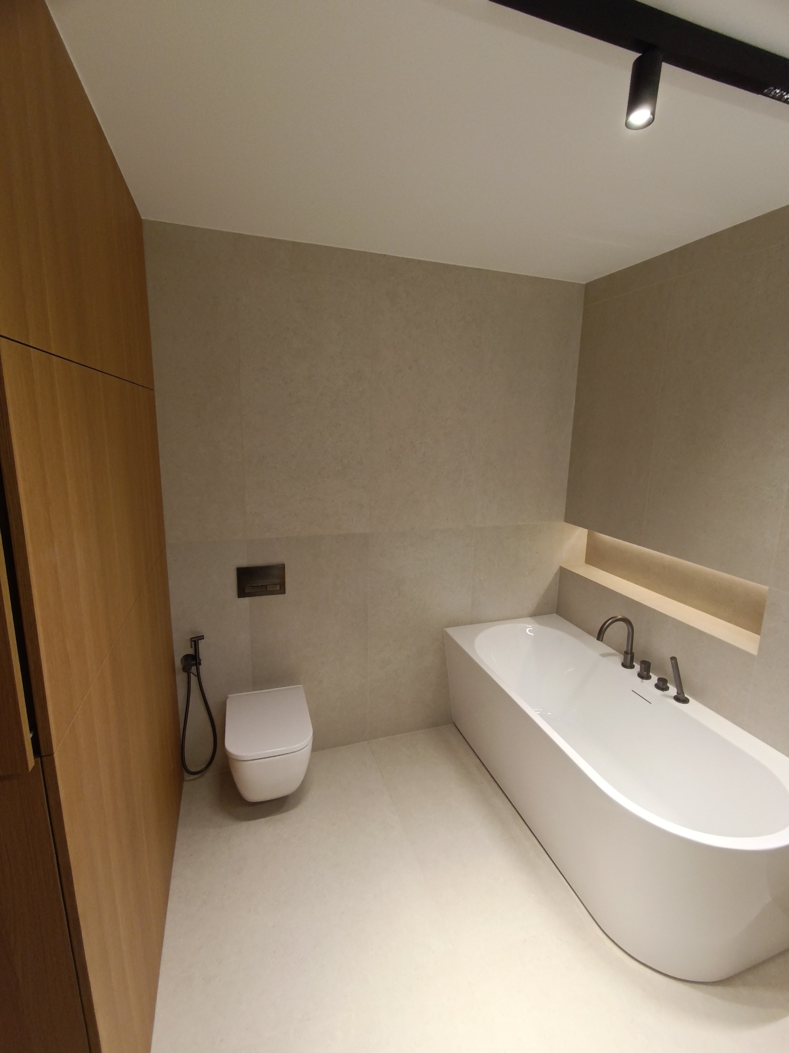 Minimalistyczna łazienka w beżach: wolnostojąca wanna, podwieszana toaleta z bidetką, drewniana zabudowa. Oświetlenie punktowe i wnękowe dodaje ciepła.