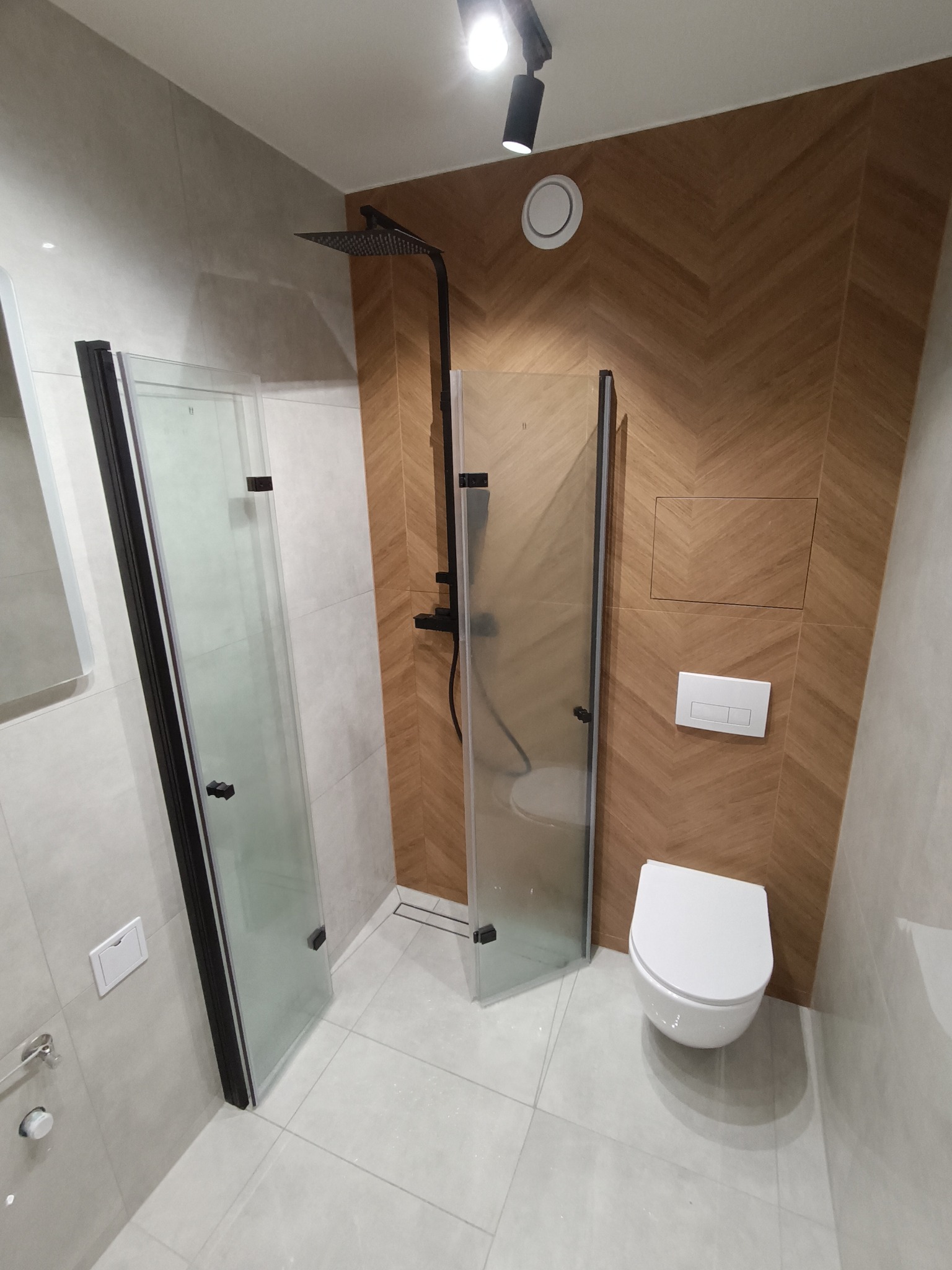 Nowoczesna łazienka z kabiną prysznicową typu walk-in z czarnymi profilami, drewnopodobną ścianą w jodełkę i podtynkową toaletą. Minimalistyczny design.