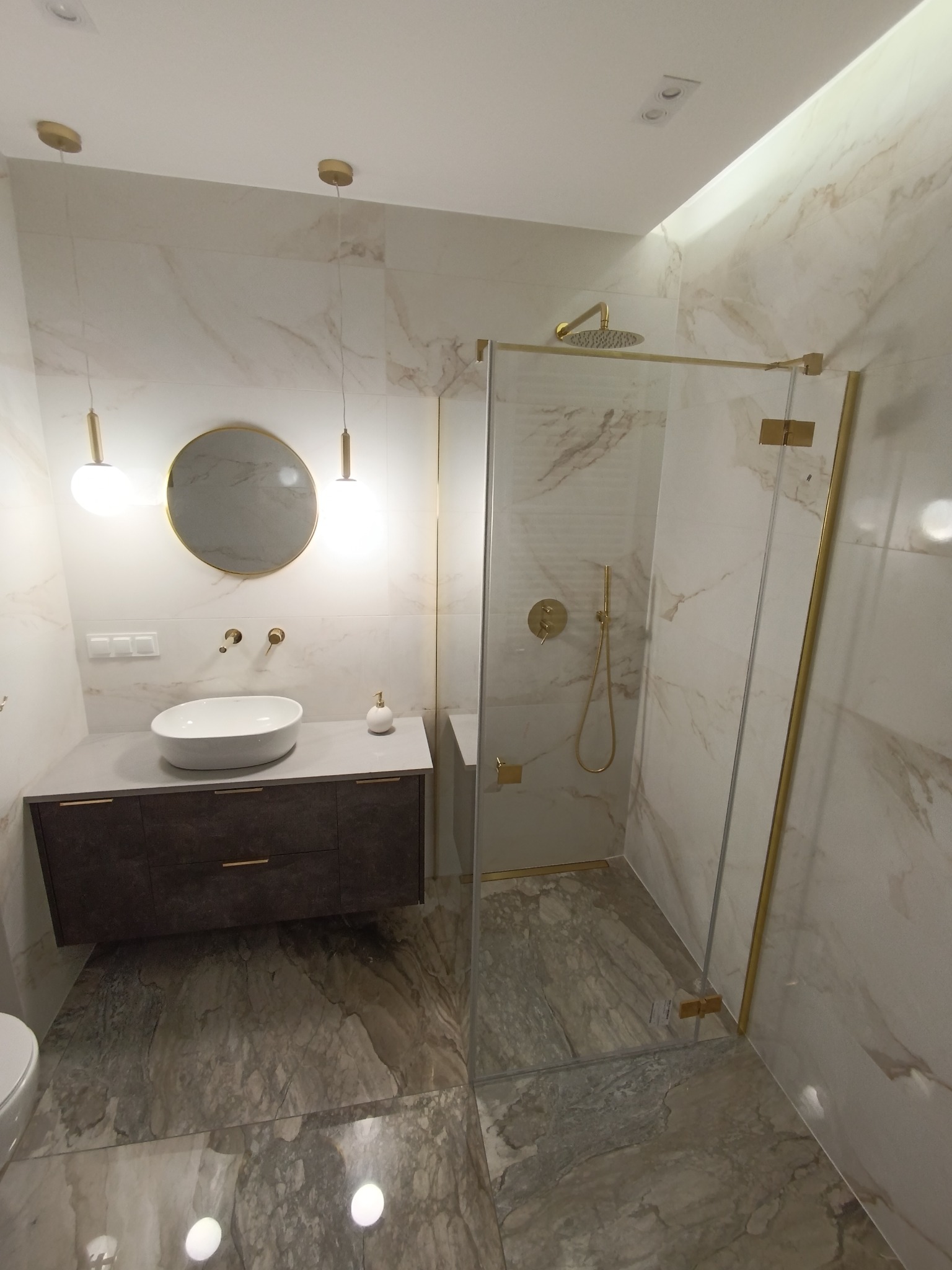 Elegancka łazienka z kabiną prysznicową typu walk-in, marmurowymi płytkami, okrągłym lustrem i złotymi akcentami. Nowoczesny design z wiszącą szafką pod umywalką.