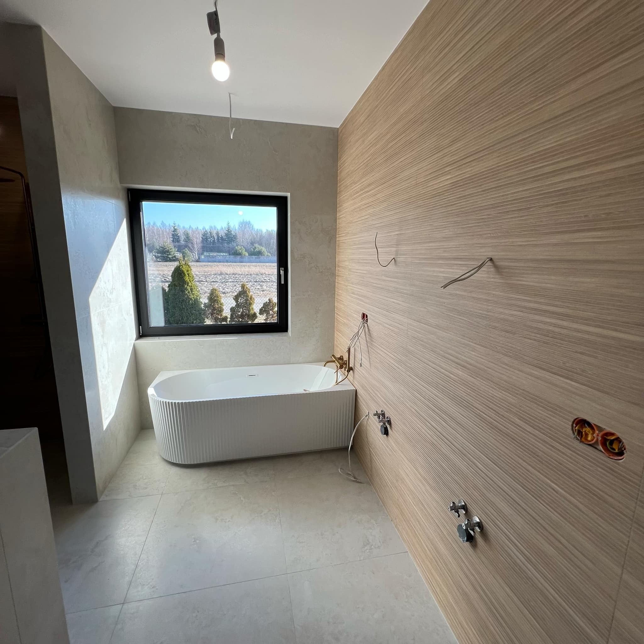 Nowoczesna łazienka w trakcie wykańczania: biała wanna, drewnopodobne panele ścienne, widoczne instalacje elektryczne i hydrauliczne. Okno z widokiem na zieleń.