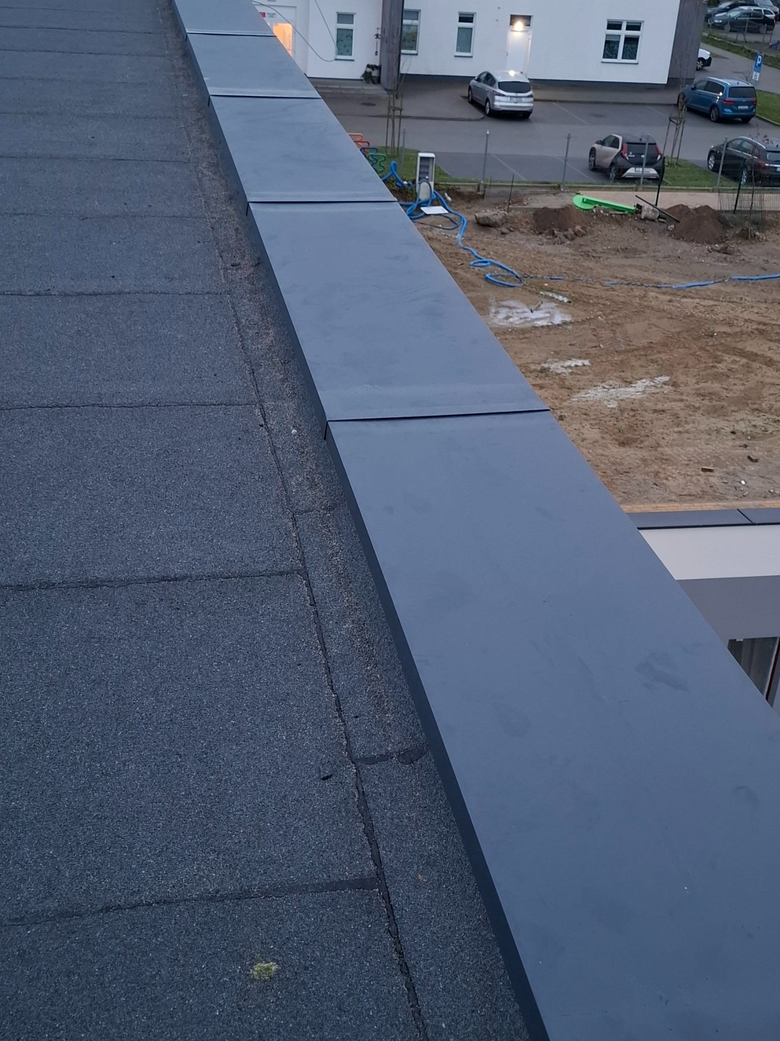 Fragment dachu pokrytego papą z widoczną obróbką blacharską w kolorze grafitowym, w tle budynek i parking.