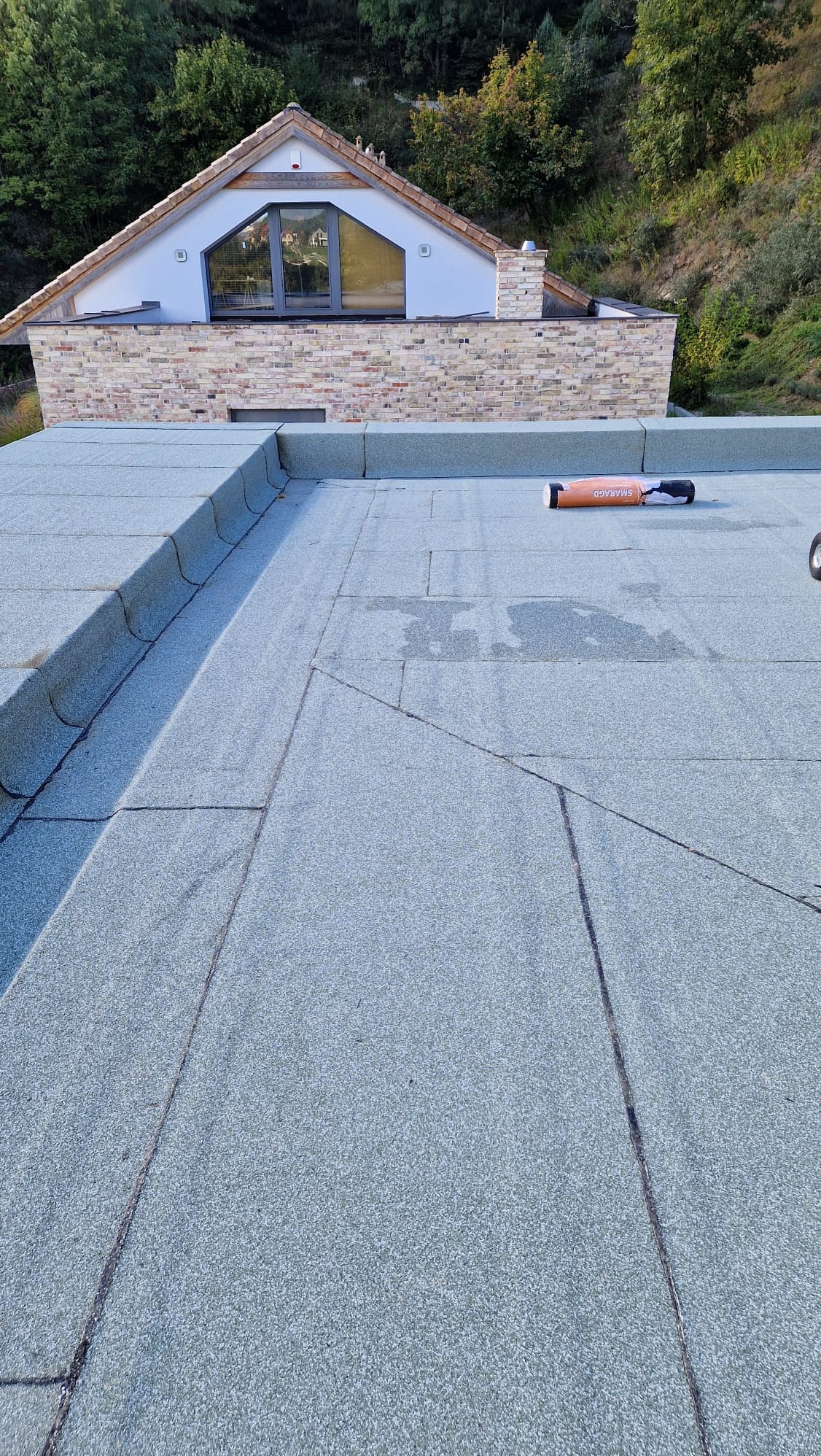 Płaski dach pokryty papą termozgrzewalną w kolorze szarym, z widocznymi łączeniami, rolka papy Smaragd na dachu, w tle budynek z cegły i las.