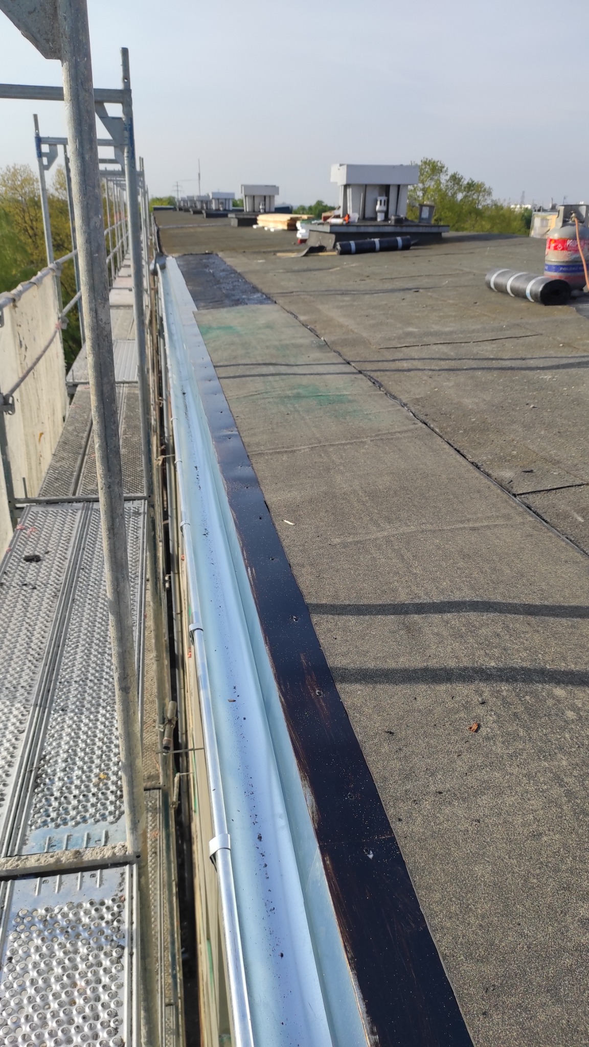 Płaski dach pokryty papą, widok z rusztowania na krawędź dachu z zamontowaną, srebrną rynną, w tle kominy wentylacyjne.