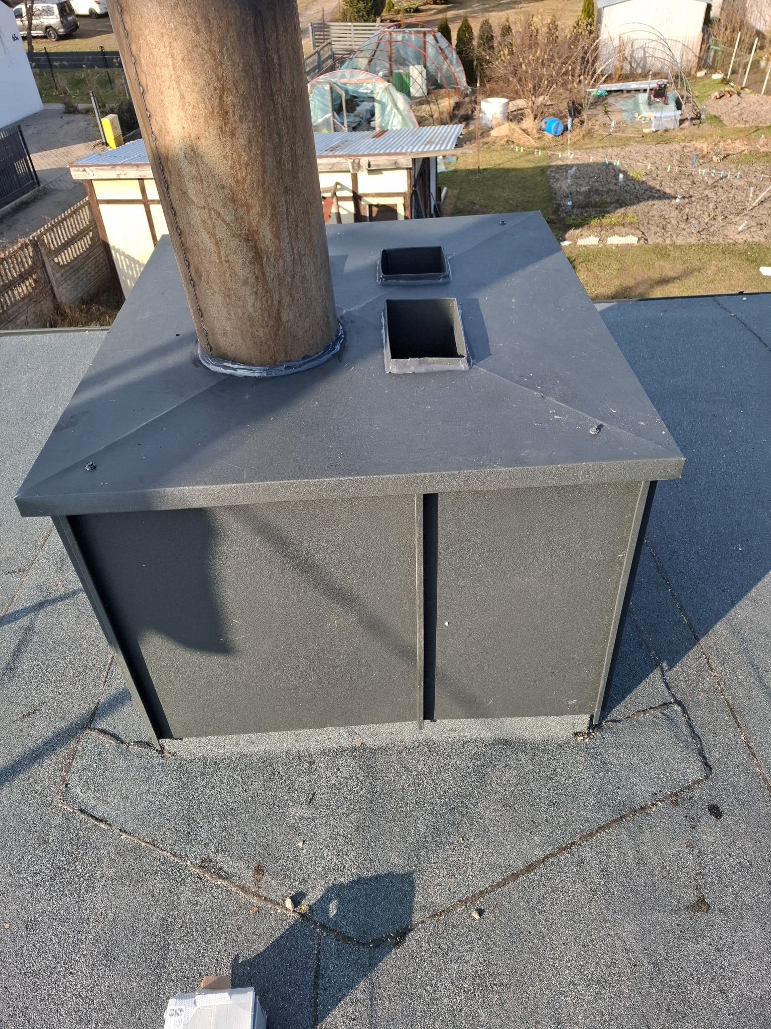 Szary komin wentylacyjny z dwoma otworami, wystający z pokrycia dachowego z papy, widok z góry z perspektywy dachu.