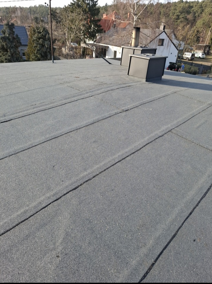 Szary dach pokryty papą termozgrzewalną z widocznymi łączeniami, dwa kominy wentylacyjne w tle, domy i drzewa w oddali.