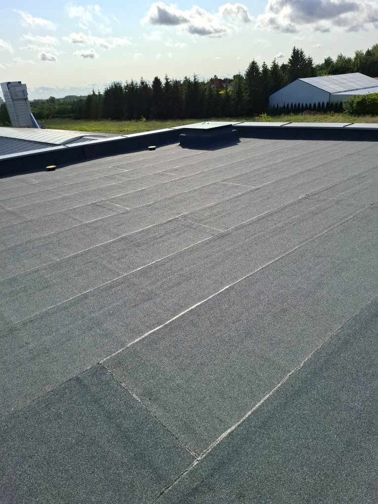 Płaski dach pokryty papą termozgrzewalną na tle zieleni i błękitnego nieba z chmurami. Widoczne łączenia papy i elementy wentylacyjne.