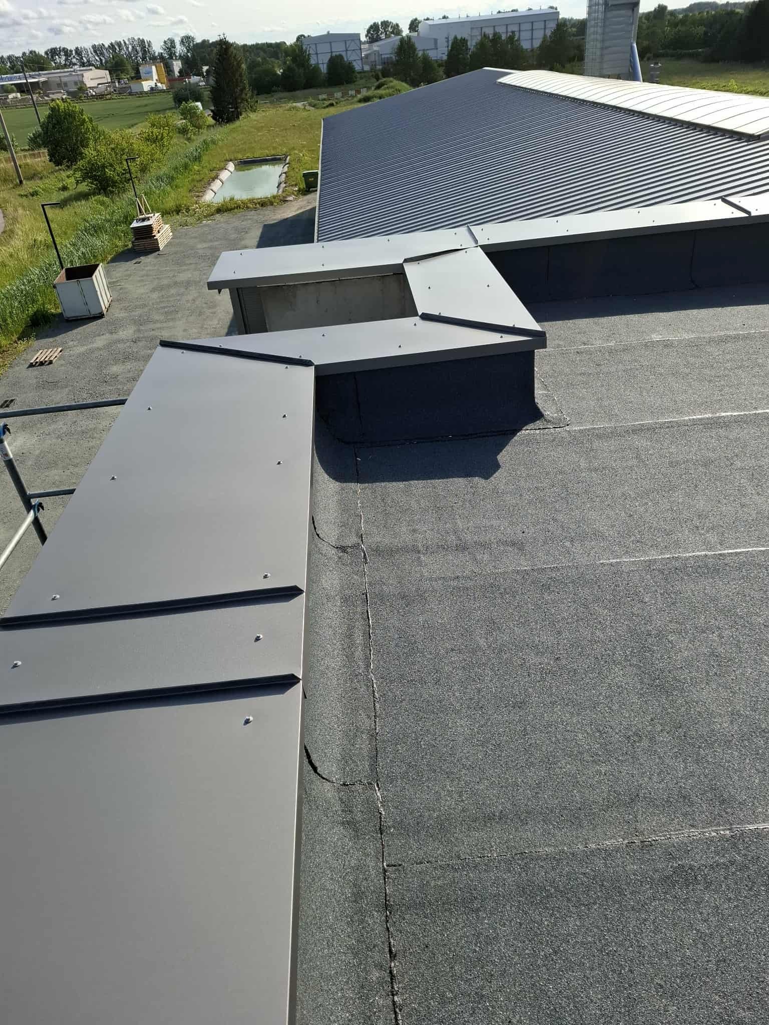 Szary dach pokryty papą z obróbkami blacharskimi. Widoczne łączenia papy i detale wykonania. W tle budynki i zieleń.