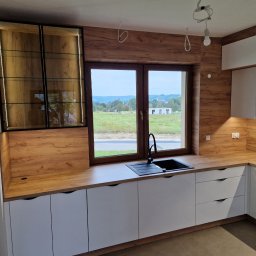 FHUP MARLON-MEBLE - Nowoczesna kuchnia z białymi szafkami i drewnianym blatem, czarny zlew pod oknem z widokiem na wiejski krajobraz, przeszklona szafka z oświetleniem LED.