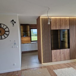 FHUP MARLON-MEBLE - Nowoczesne wnętrze z elementami drewna: ażurowa ścianka działowa z podświetleniem LED, częściowo widoczna zabudowa kuchenna z białymi frontami i drewnianymi szafkami wiszącymi, duży zegar ścienny...