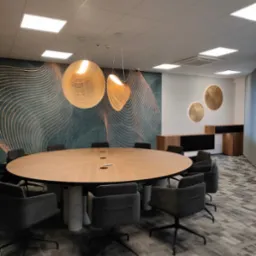 Owalny stół konferencyjny z jasnego drewna z wbudowanymi gniazdami, otoczony szarymi krzesłami na kółkach, podwieszane lampy z drewnianych listewek i dekoracyjna tapeta w tle.