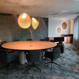 FHUP MARLON-MEBLE - Owalny stół konferencyjny z drewnianym blatem, wsparty na cylindrycznych marmurowych podstawach, otoczony szarymi krzesłami biurowymi na kółkach, oświetlony wiszącymi lampami z ażurowymi kloszami...