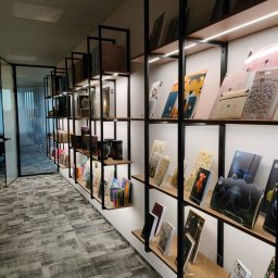 FHUP MARLON-MEBLE - Długa, wąska półka z czarnymi metalowymi ramami i drewnianymi półkami, oświetlona listwą LED, wypełniona różnorodnymi artykułami biurowymi i książkami, widok z perspektywy korytarza biurowego...