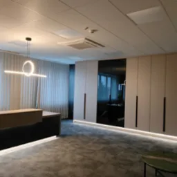 Nowoczesne biuro z recepcją: minimalistyczne szafy wnękowe z czarnymi, pionowymi uchwytami, podświetlane od dołu, oraz recepcja z czarnym frontem i beżowym blatem, oświetlona okrągłą lampą wiszącą.