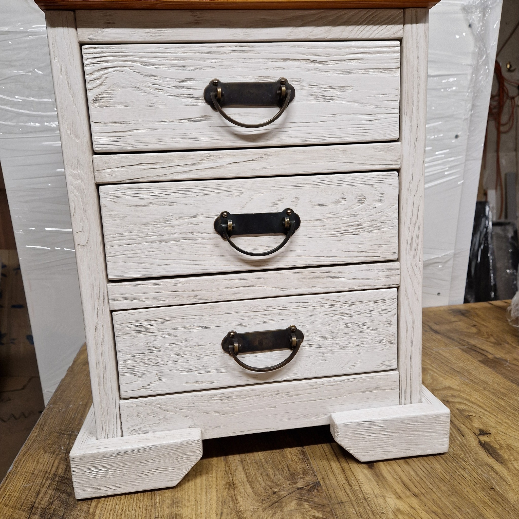 Drewniana komoda z trzema szufladami w stylu rustykalnym, bielona z brązowym blatem i metalowymi uchwytami, widoczna struktura drewna.