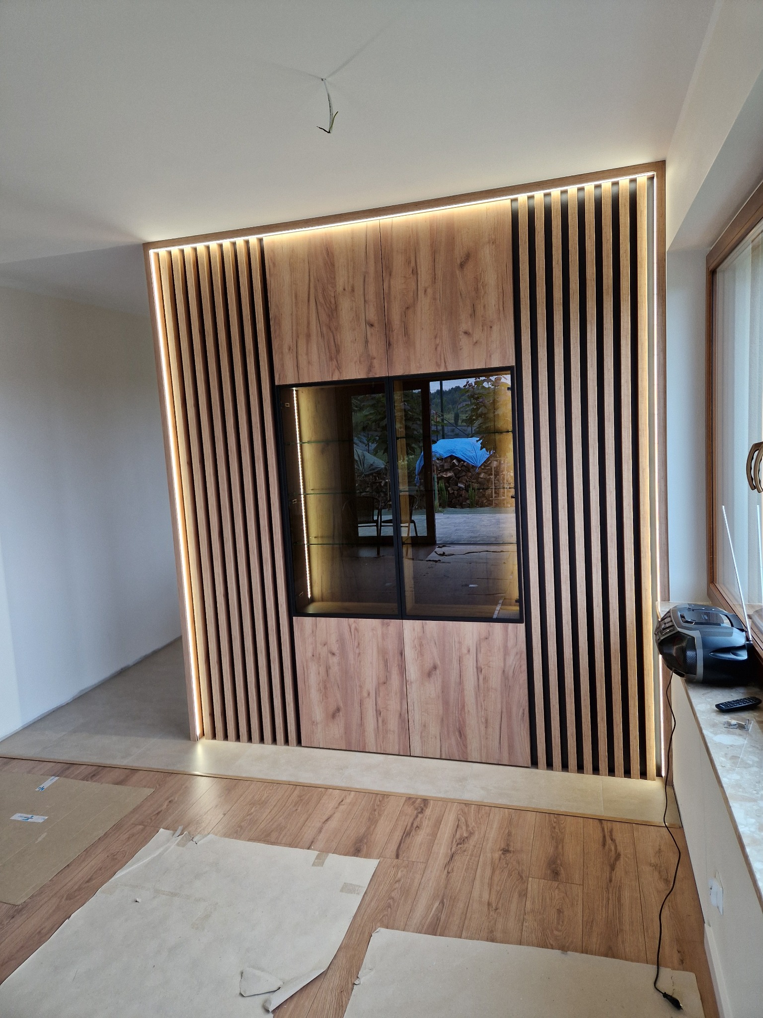 Drewniana zabudowa ściany z pionowymi lamelami i podświetleniem LED, wbudowana przeszklona witryna z widokiem na zewnątrz, częściowo zakryta podłoga.