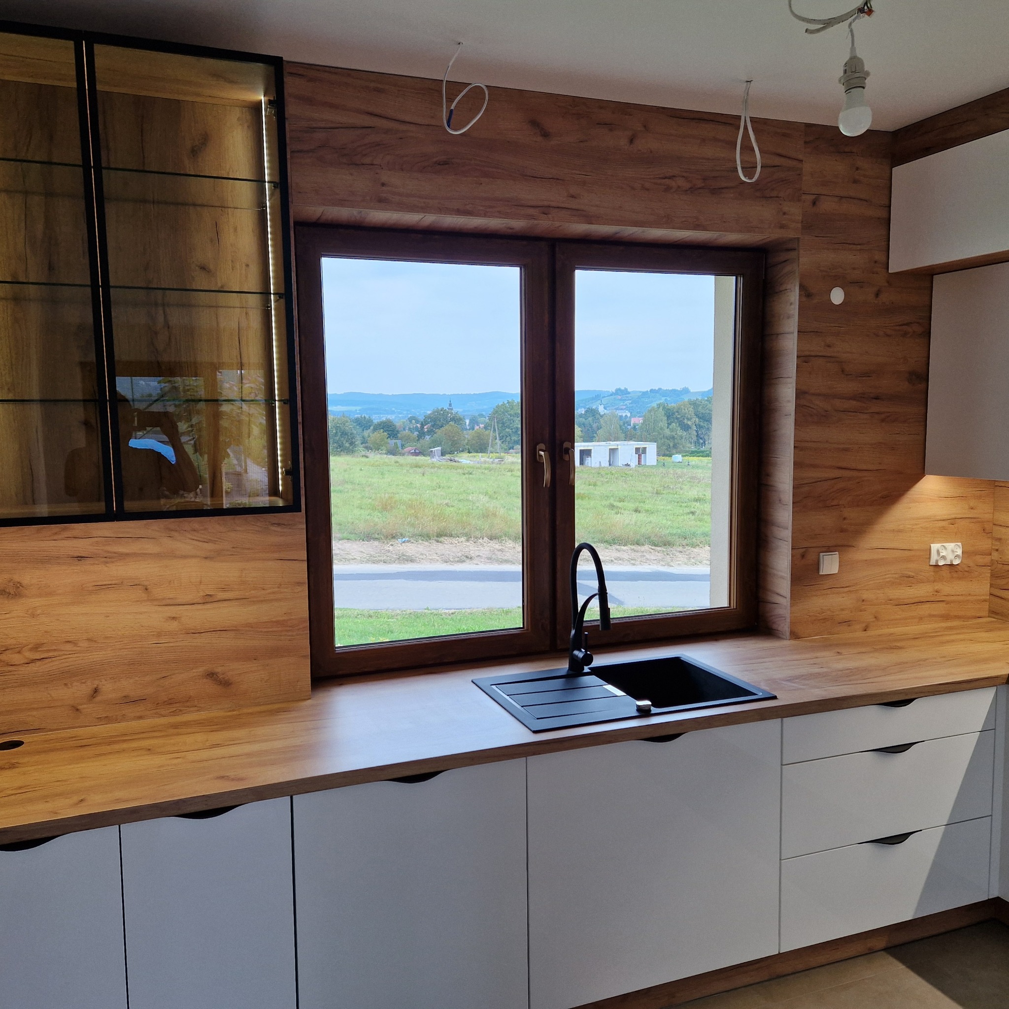 Nowoczesna kuchnia z białymi szafkami i drewnianym blatem, czarny zlew pod oknem z widokiem na zielone pola i budynek w budowie, wisząca szafka z podświetleniem.