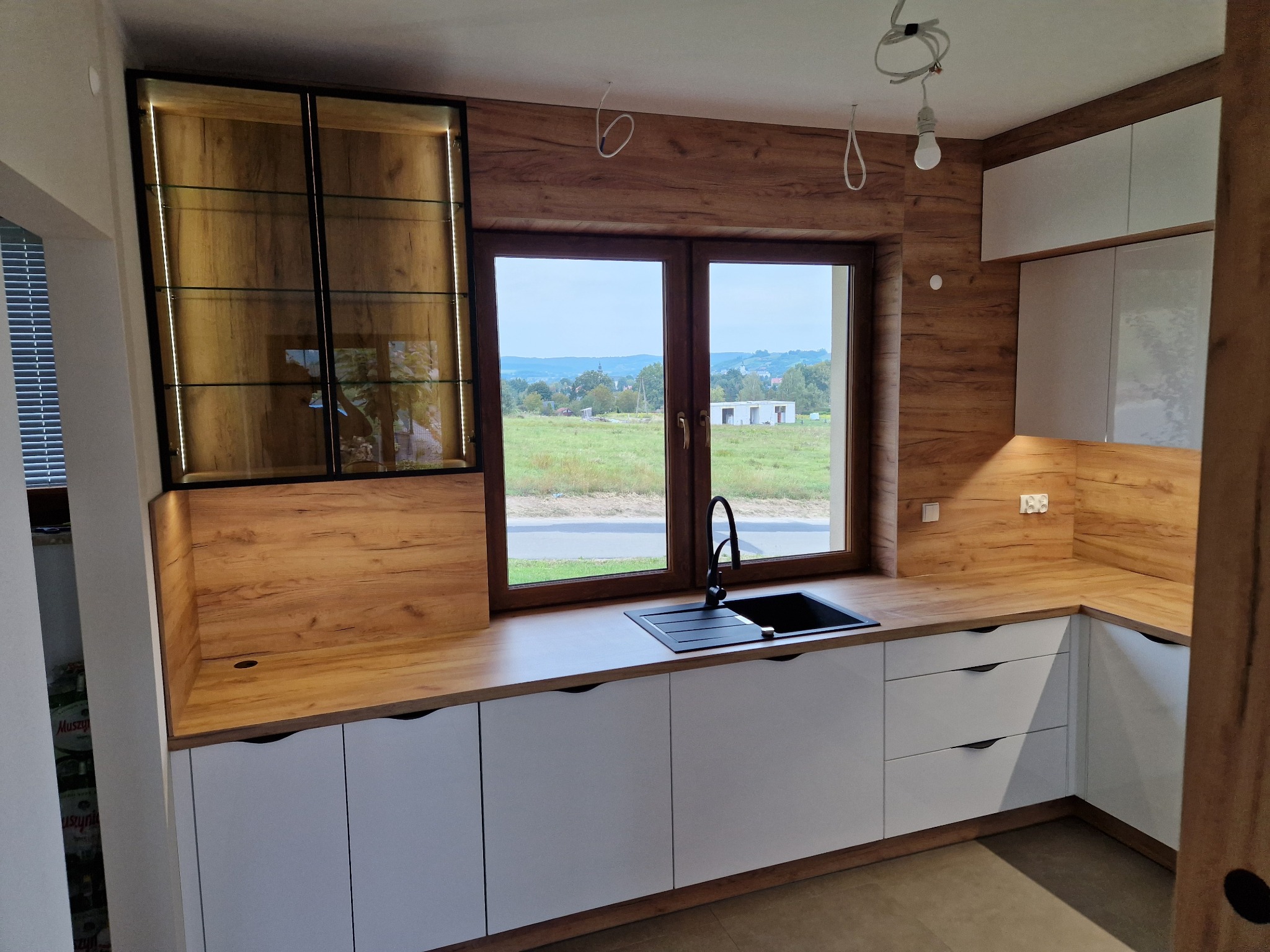 Nowoczesna kuchnia z białymi szafkami i drewnianym blatem, czarny zlew pod oknem z widokiem na wiejski krajobraz, przeszklona szafka z oświetleniem LED.