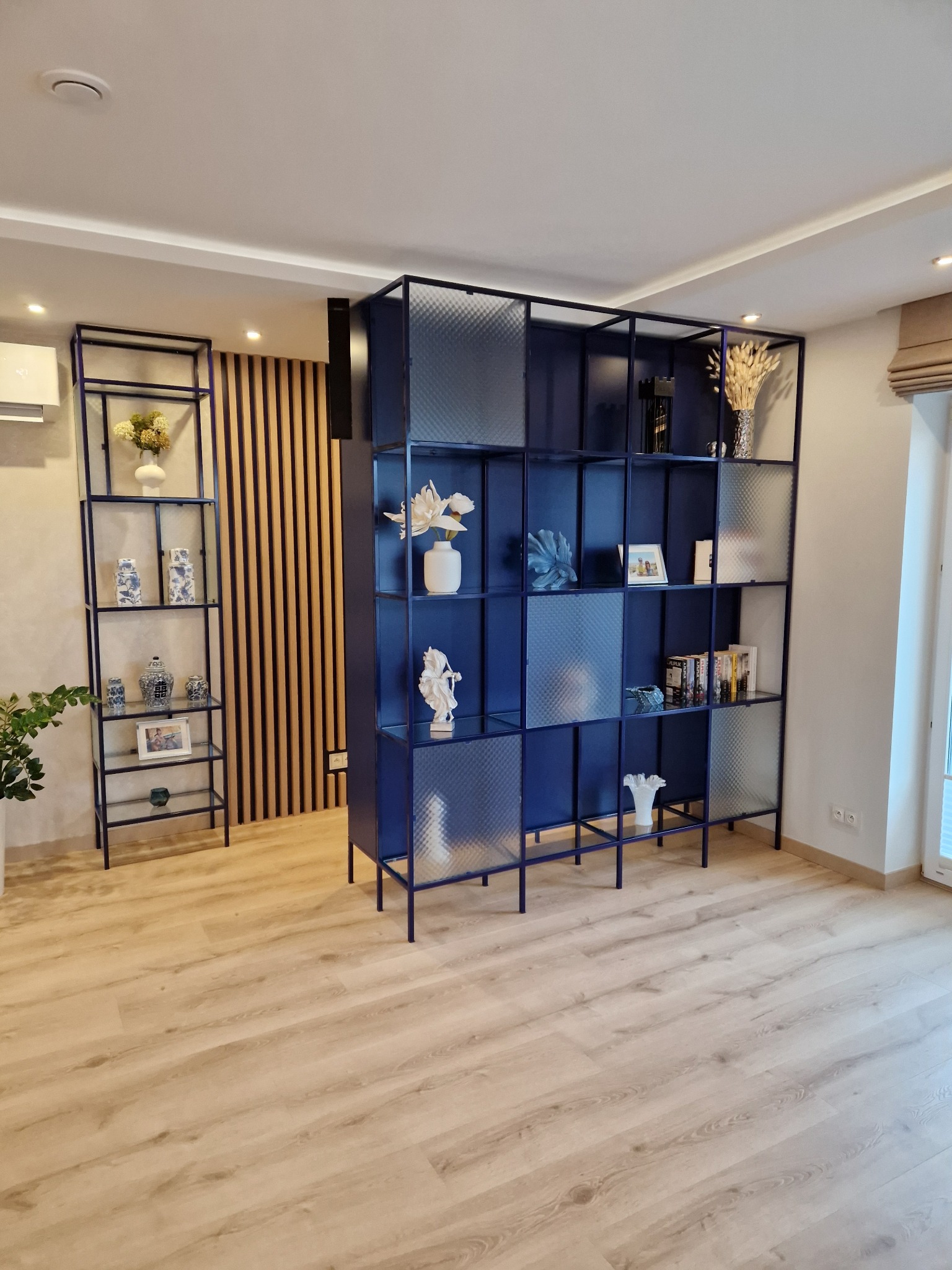 Niebieski regał modułowy z przeszkleniami w kratkę, wypełniony dekoracjami, w jasnym salonie z drewnianą podłogą i ścianą z drewnianych paneli.