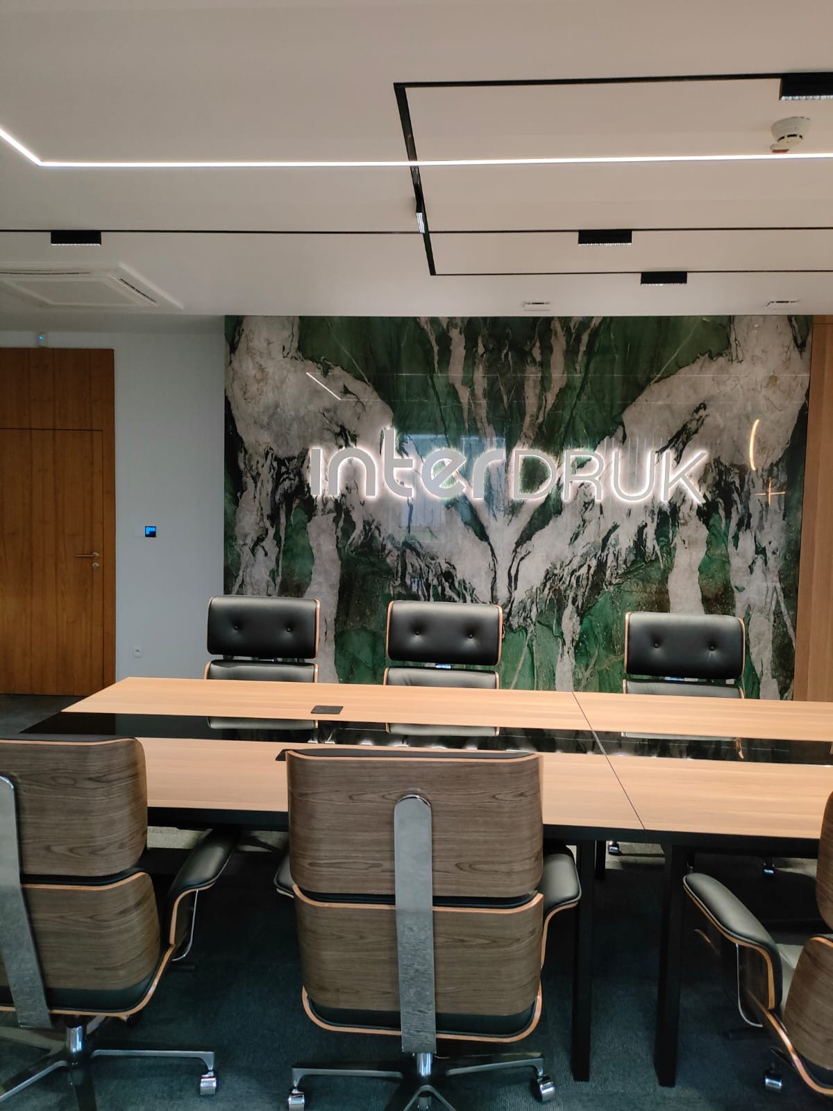 Sala konferencyjna z długim stołem, czarnymi krzesłami i ścianą z zielonego marmuru z podświetlonym neonem 'interDRUK'.