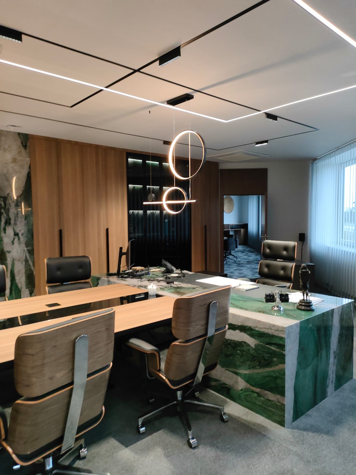 Nowoczesne biuro z długim stołem konferencyjnym, krzesłami z drewnianym wykończeniem, marmurowym blatem i designerskim oświetleniem sufitowym.