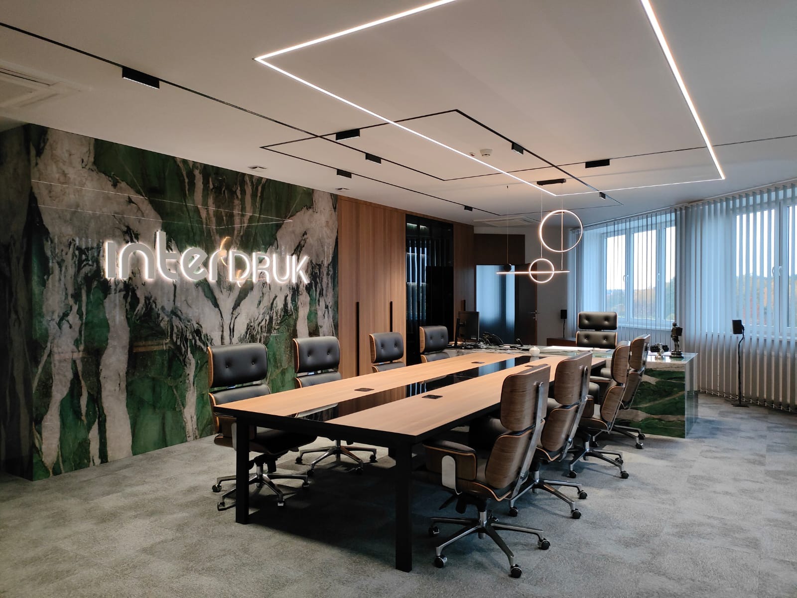 Sala konferencyjna z długim drewnianym stołem, czarnymi krzesłami i neonowym logo firmy na ścianie z zielonego marmuru, oświetlona nowoczesnymi lampami sufitowymi.