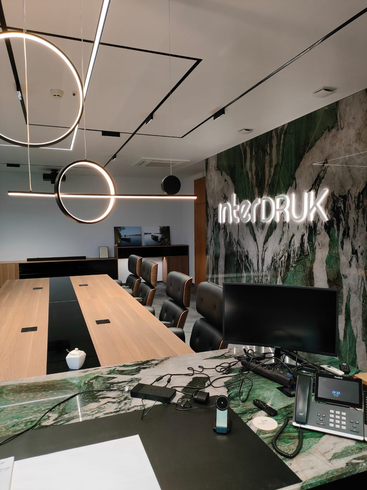 Sala konferencyjna z dużym stołem z drewnianym blatem i czarnym pasem pośrodku, czarne skórzane krzesła, marmurowa ściana z podświetlanym napisem 'interDRUK', nowoczesne oświetlenie sufitowe.