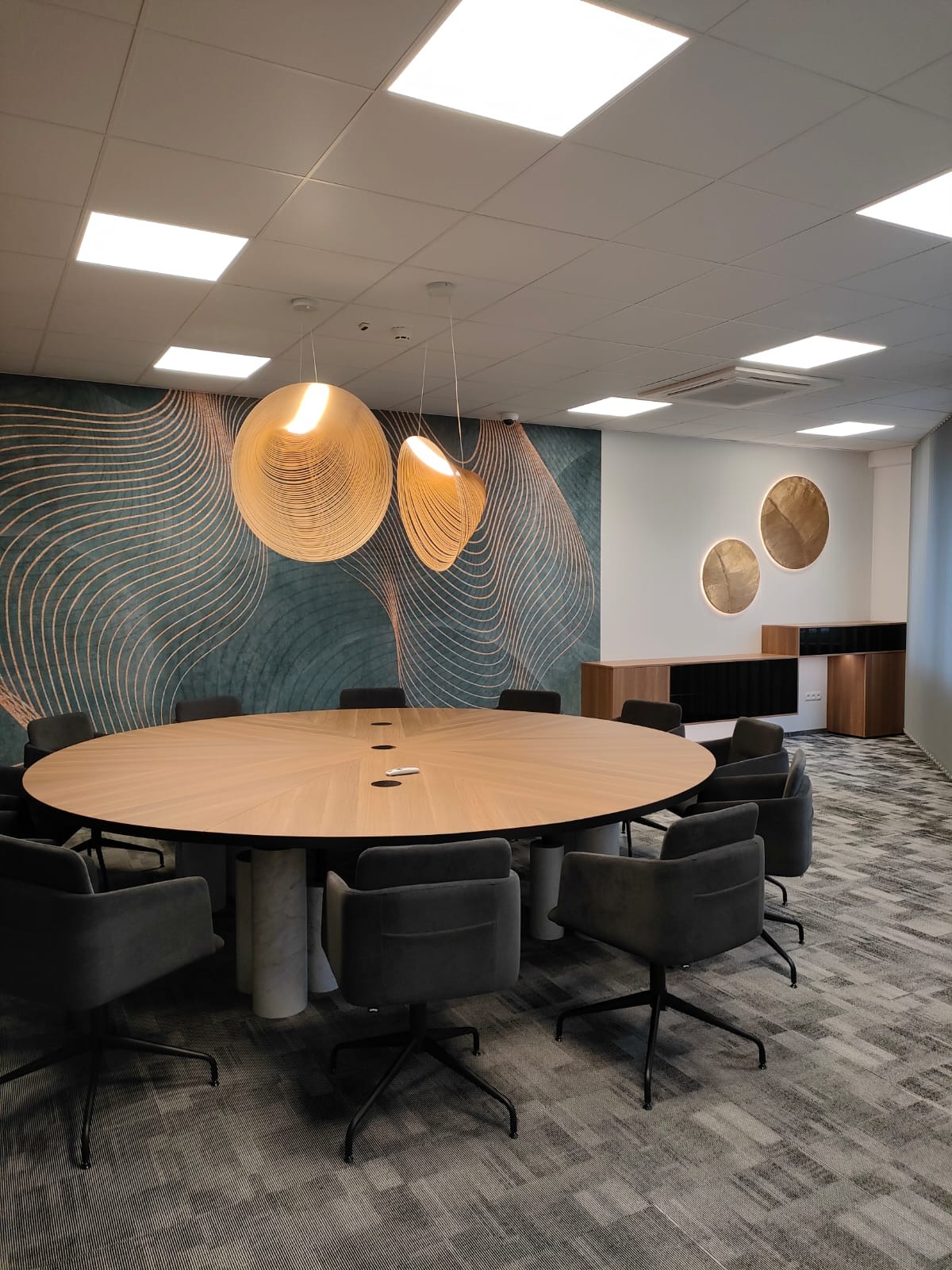 Owalny stół konferencyjny z jasnego drewna z wbudowanymi gniazdami, otoczony szarymi krzesłami na kółkach, podwieszane lampy z drewnianych listewek i dekoracyjna tapeta w tle.