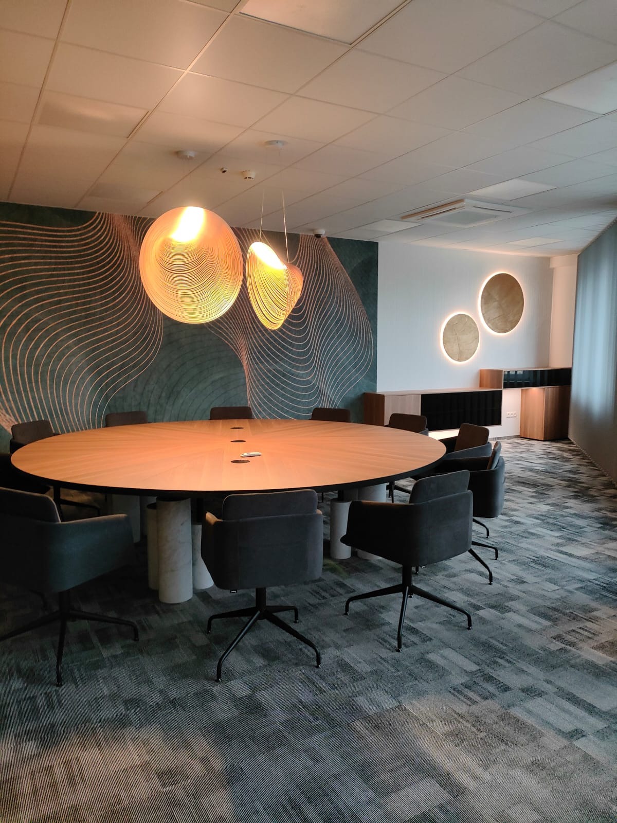 Owalny stół konferencyjny z drewnianym blatem, wsparty na cylindrycznych marmurowych podstawach, otoczony szarymi krzesłami biurowymi na kółkach, oświetlony wiszącymi lampami z ażurowymi kloszami...