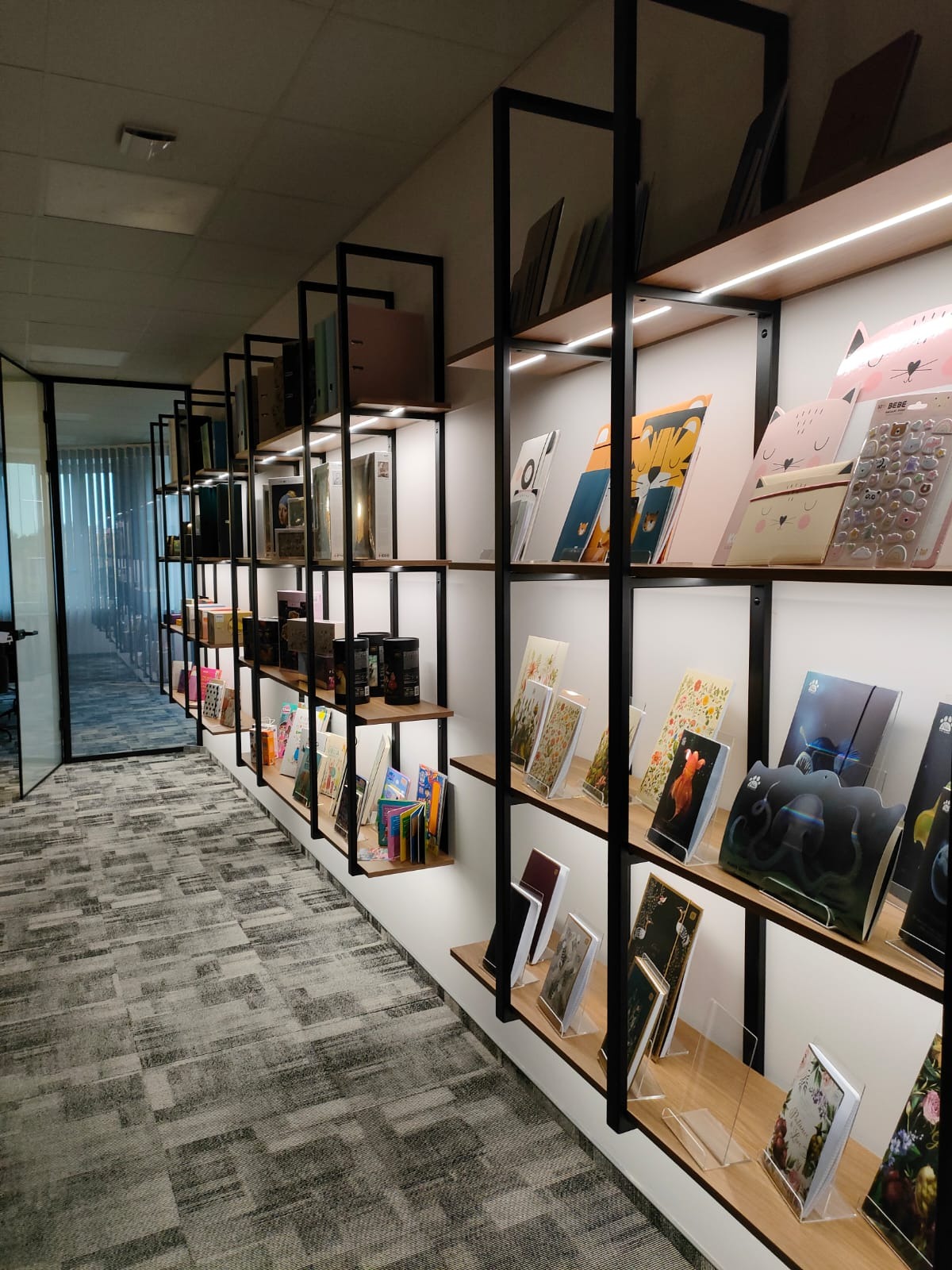 Długa, wąska półka z czarnymi metalowymi ramami i drewnianymi półkami, oświetlona listwą LED, wypełniona różnorodnymi artykułami biurowymi i książkami, widok z perspektywy korytarza biurowego...