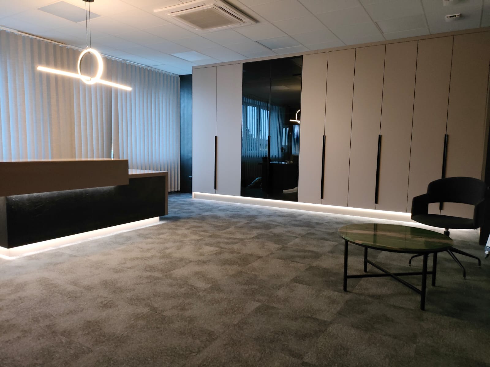 Nowoczesne biuro z recepcją, szafą wnękową z lustrzanymi drzwiami i podświetleniem LED, okrągłym stolikiem kawowym i fotelem na tle zasłon.