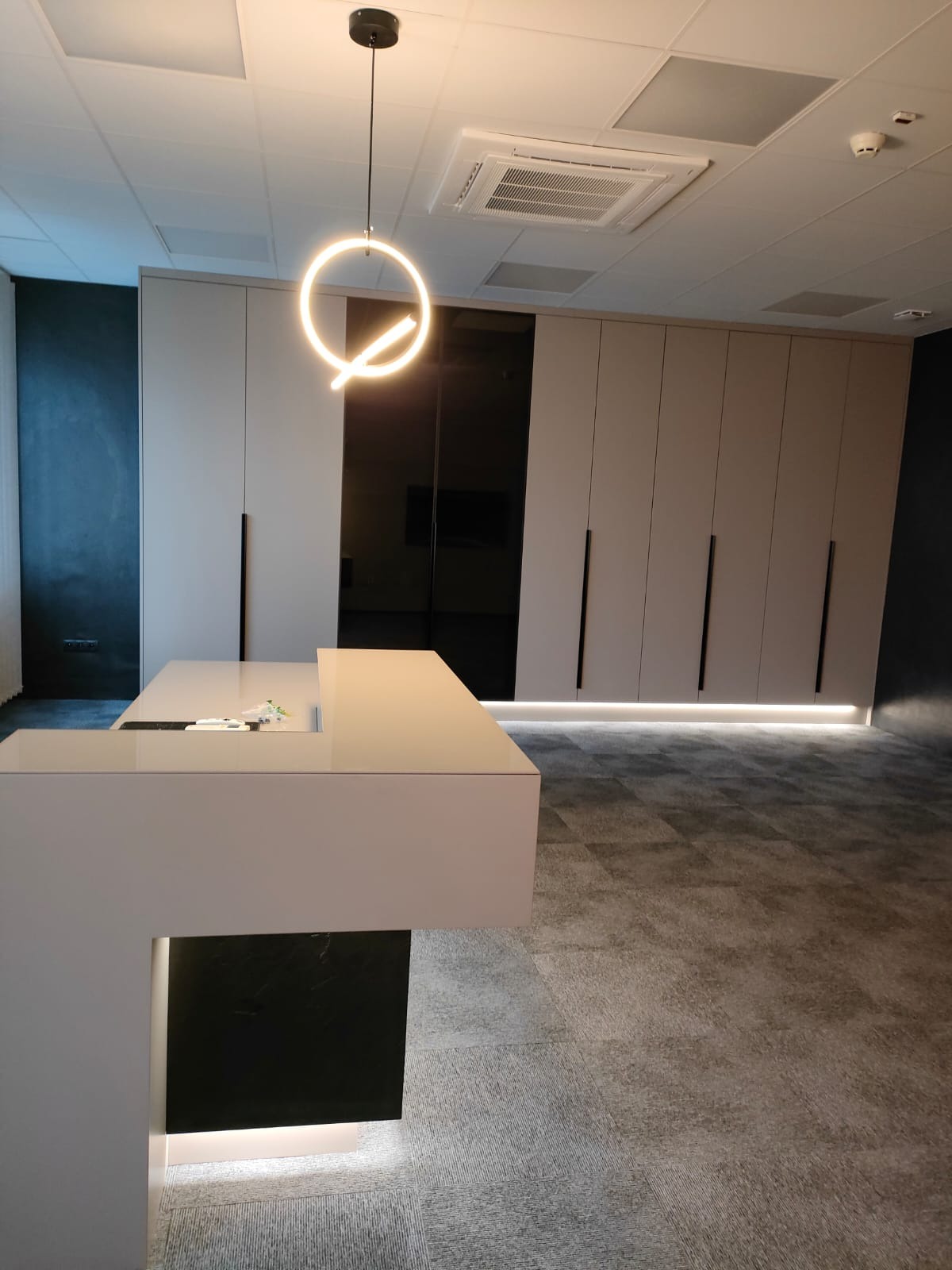 Nowoczesne biuro z szafami w zabudowie o jasnych frontach, podświetleniem LED u dołu i czarnymi uchwytami, oraz recepcją z czarnym panelem i okrągłą lampą wiszącą.