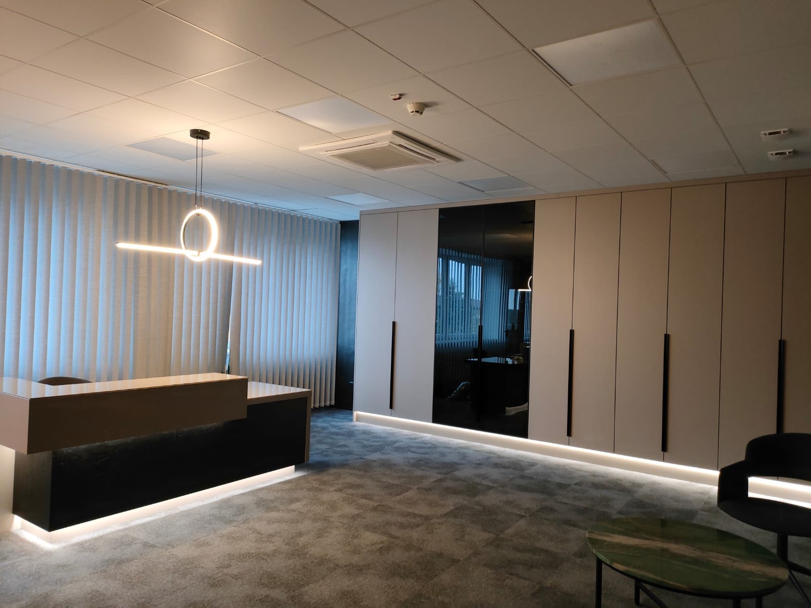 Nowoczesne biuro z recepcją: minimalistyczne szafy wnękowe z czarnymi, pionowymi uchwytami, podświetlane od dołu, oraz recepcja z czarnym frontem i beżowym blatem, oświetlona okrągłą lampą wiszącą.