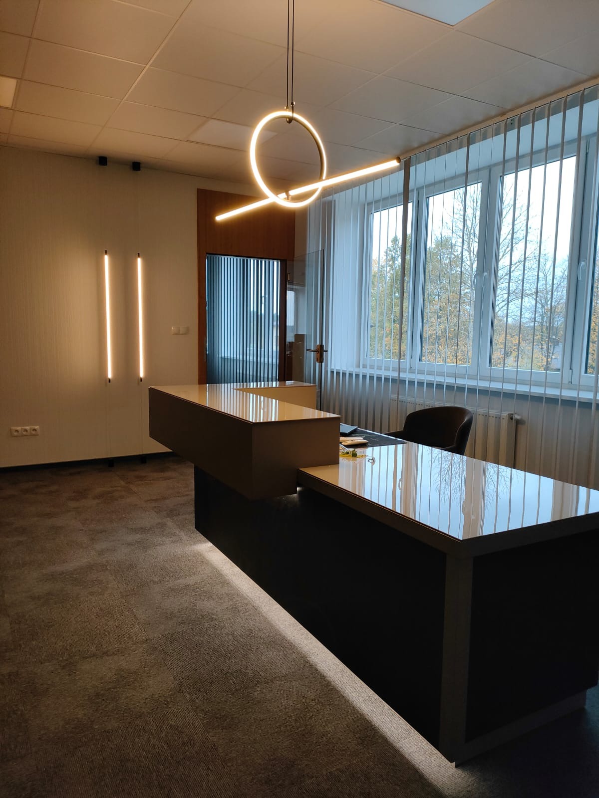 Nowoczesne biuro z biurkiem na wymiar, oświetleniem LED w kształcie okręgu i pionowymi lampami ściennymi, widok na zewnątrz przez żaluzje.