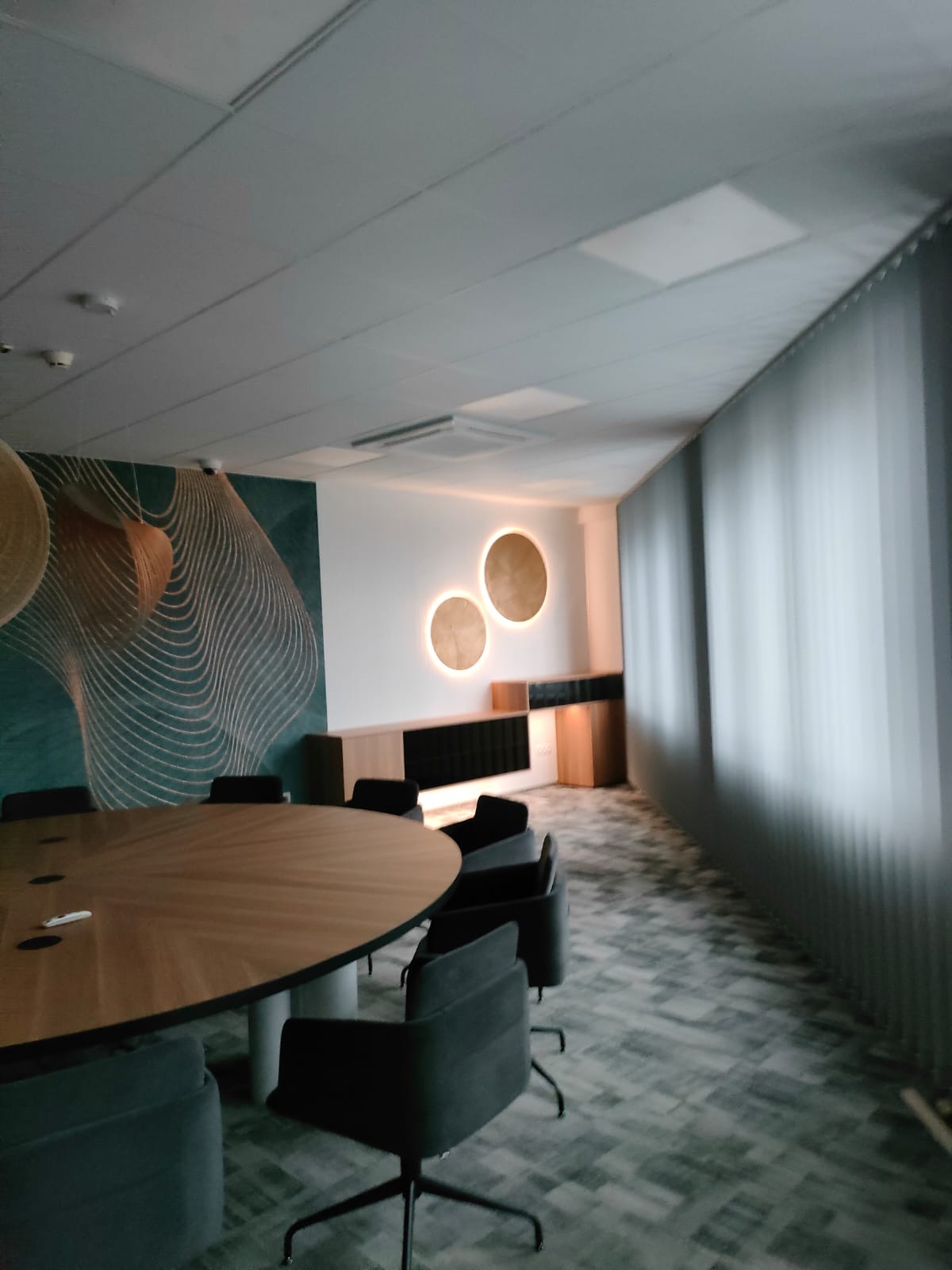 Nowoczesna sala konferencyjna z owalnym stołem z jasnego drewna, krzesłami na kółkach w kolorze grafitowym, zabudową meblową podświetloną okrągłymi lampami na ścianie oraz ścianą z abstrakcyjnym...