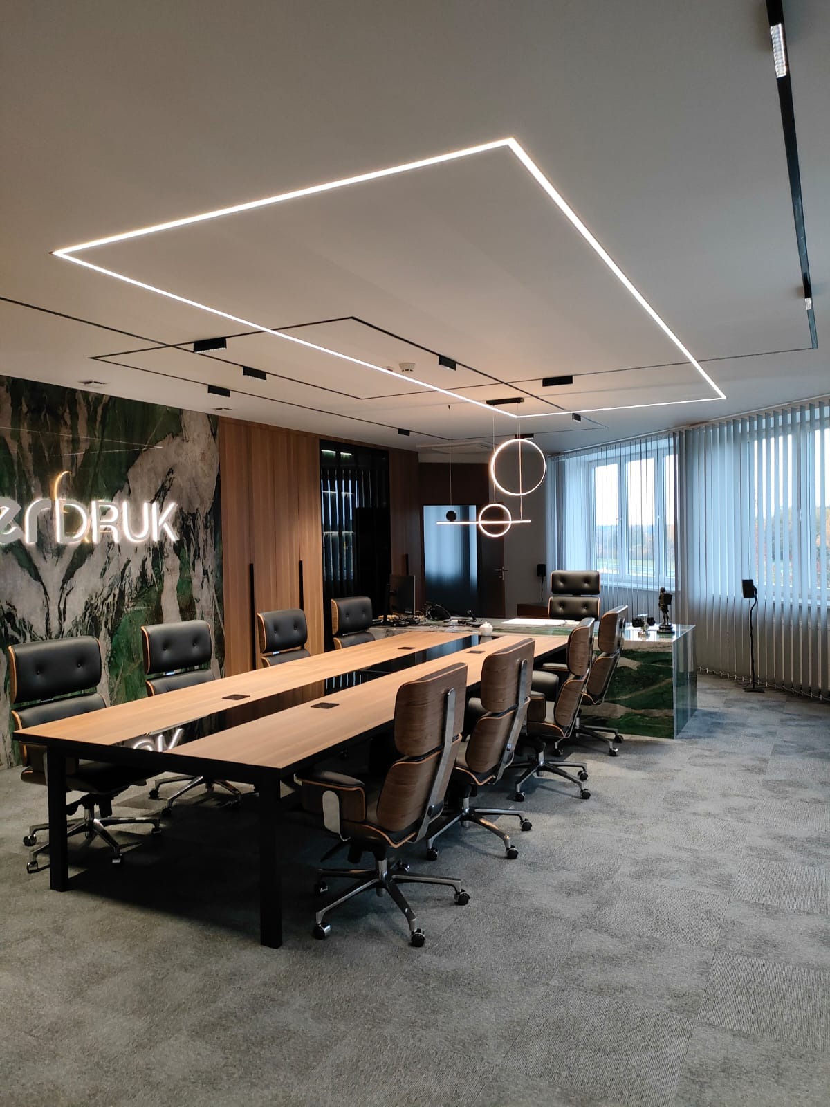 Nowoczesna sala konferencyjna z dużym drewnianym stołem, czarnymi krzesłami, marmurową ścianą z neonowym napisem i designerskim oświetleniem sufitowym.