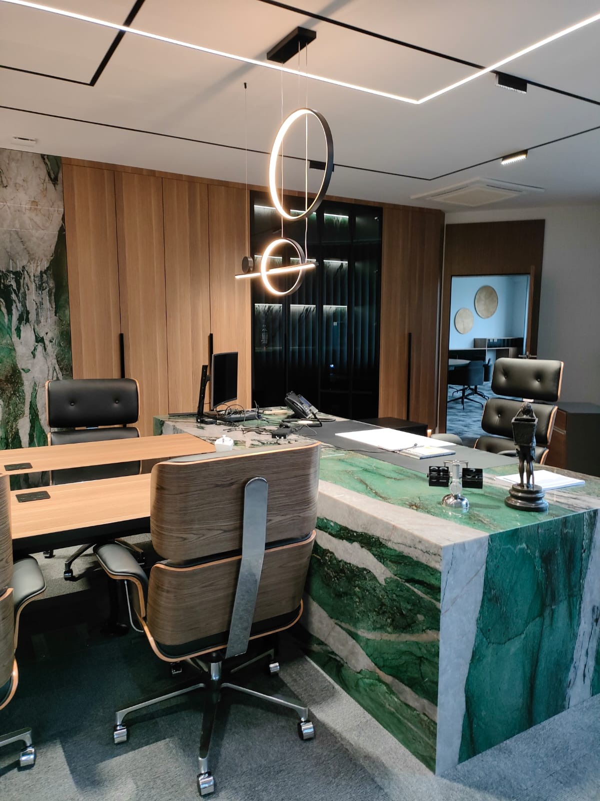 Nowoczesne biuro z dużym biurkiem z zielonego marmuru, drewnianymi szafami w tle i designerskim oświetleniem sufitowym.