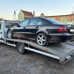 Stanisław Bróździński - Czarny Mercedes na srebrnej lawecie z najazdami, zabezpieczony czerwonymi pasami. W tle budynki i drzewa. Usługa transportu pojazdów.