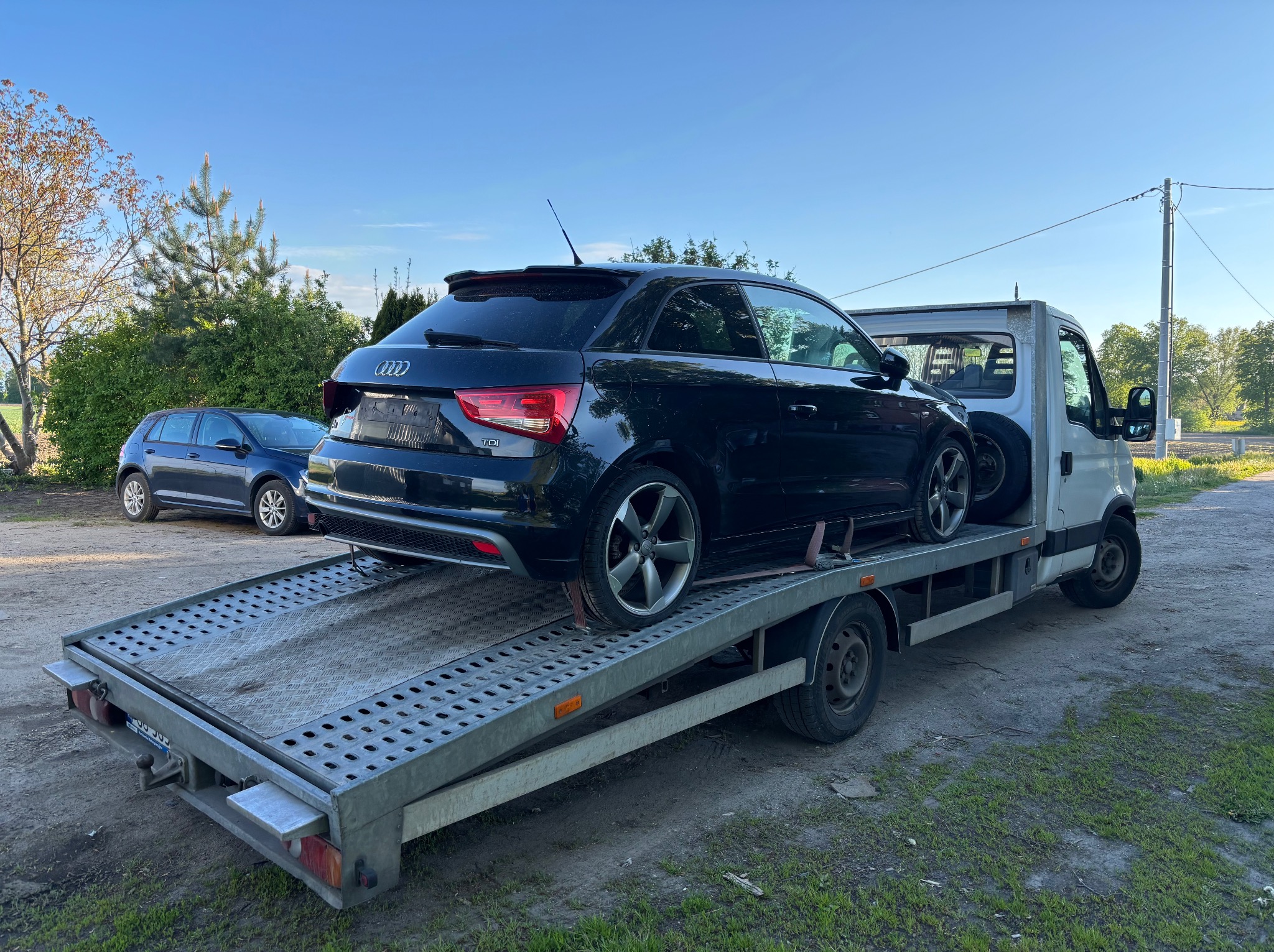 Czarny hatchback Audi A1 na platformie lawety, widok z tyłu, z boku. W tle zaparkowany granatowy Volkswagen Golf. Błękitne niebo, drzewa.