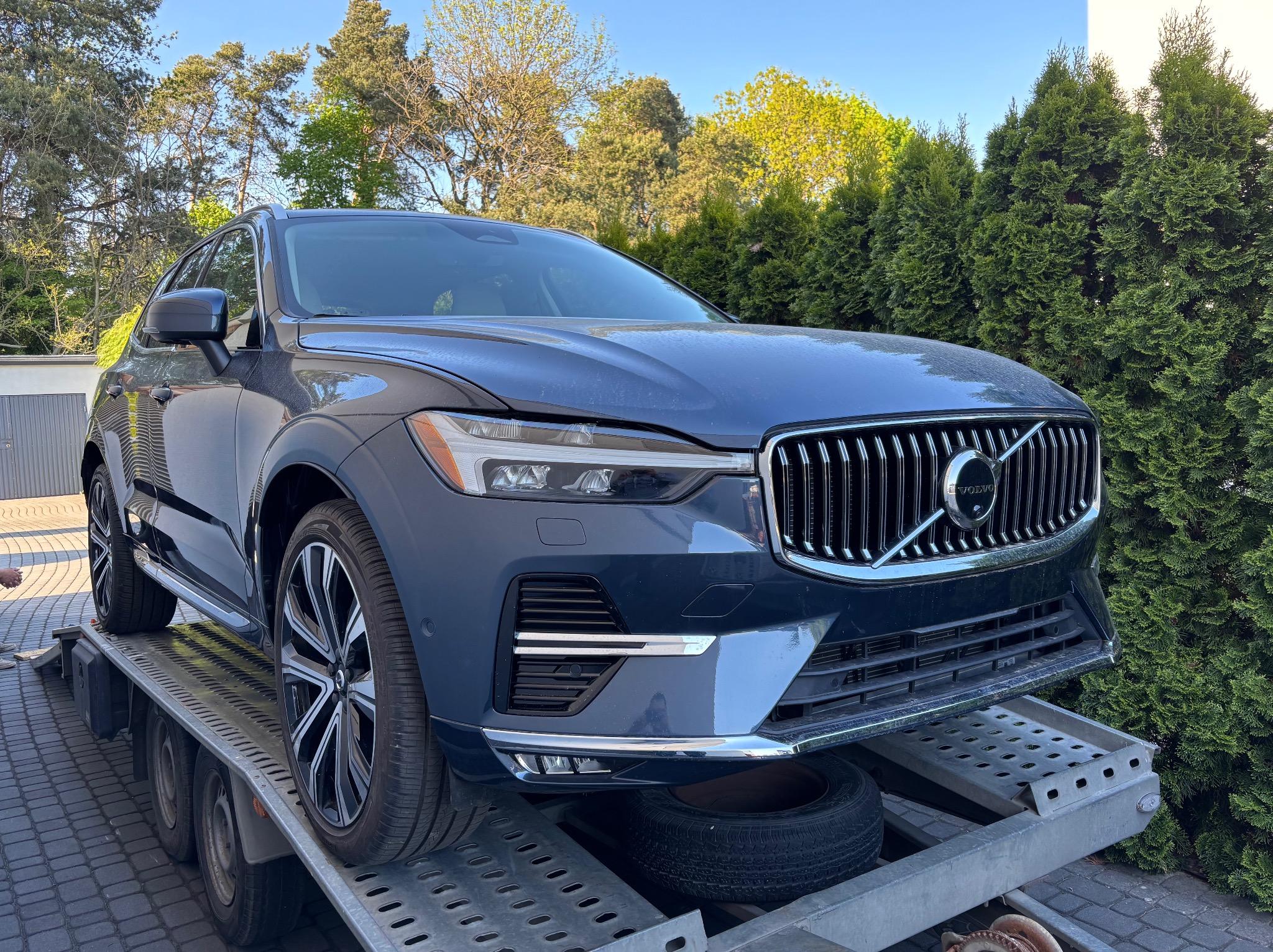 Granatowy Volvo XC60 na lawecie, z widocznym kołem zapasowym pod spodem, na tle zielonych drzew i błękitnego nieba. Usługa lawetowania.